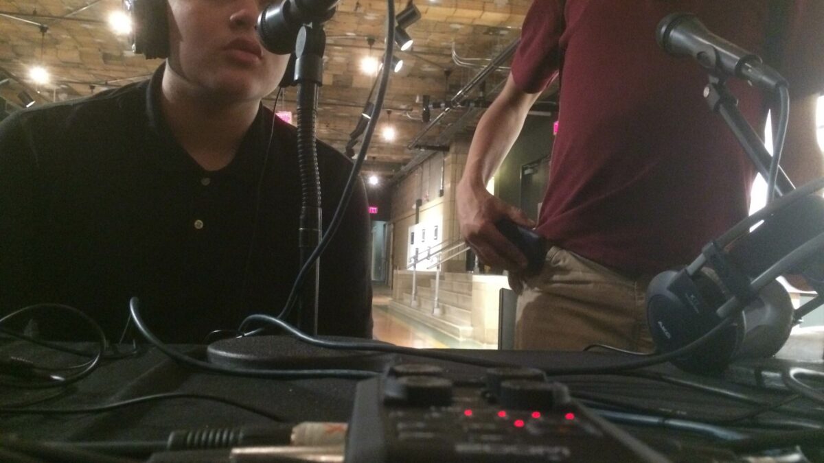 A teen wearing headphones speaking into a microphone connected to a portable recorder and an individual standing nearby