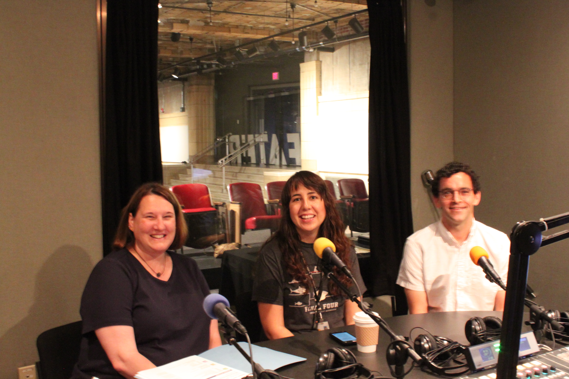Three adults smiling sitting behind microphones in a radio studio
