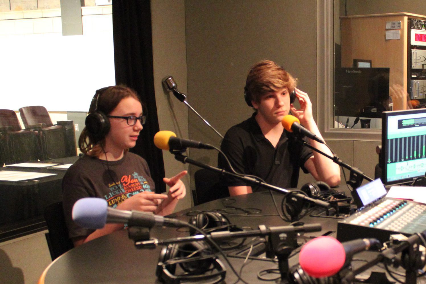 Two teens wearing headphones speaking into microphones in a radio studio