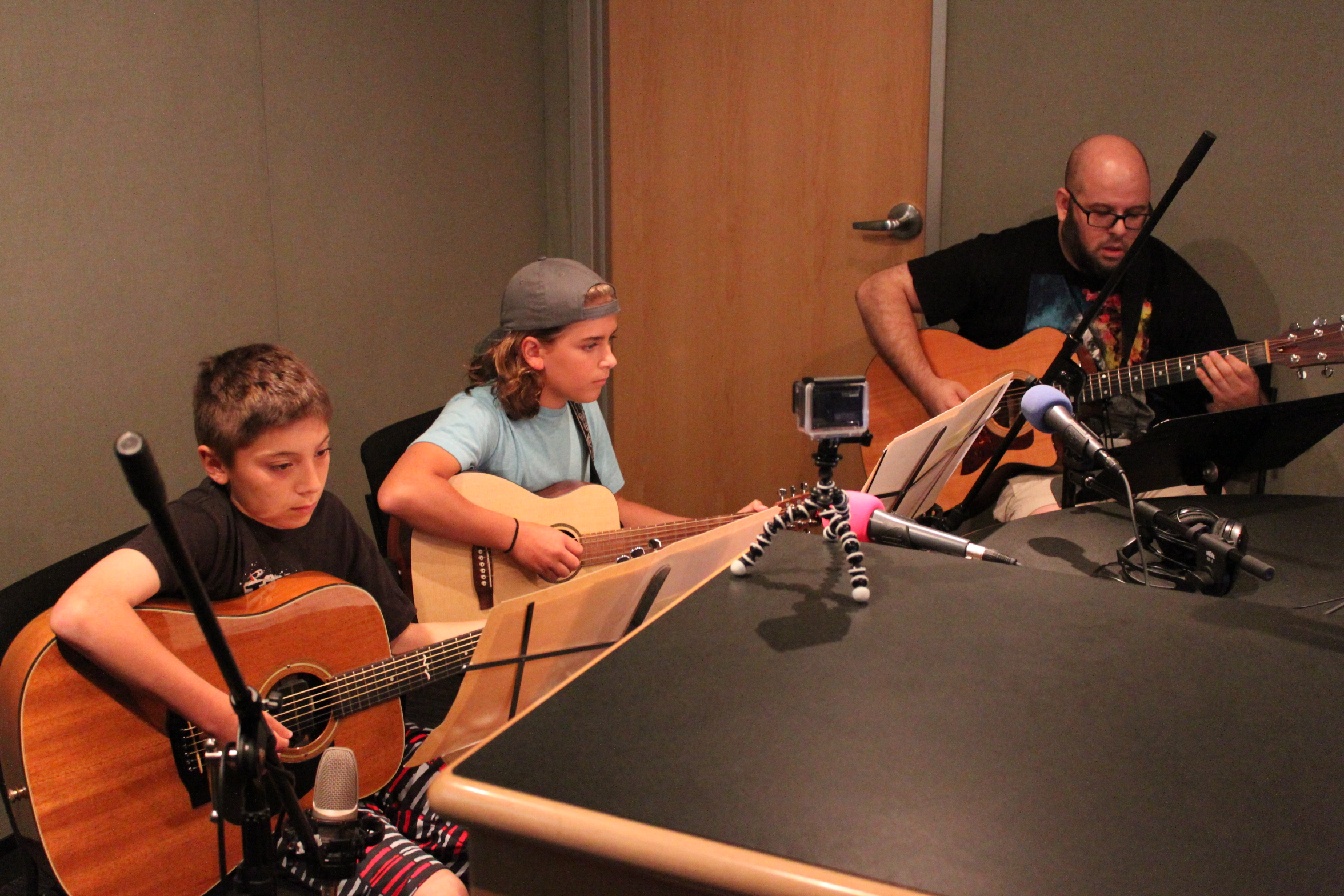 Two youth and an adult playing acoustic guitars looking at sheet music on music stands in a radio studio