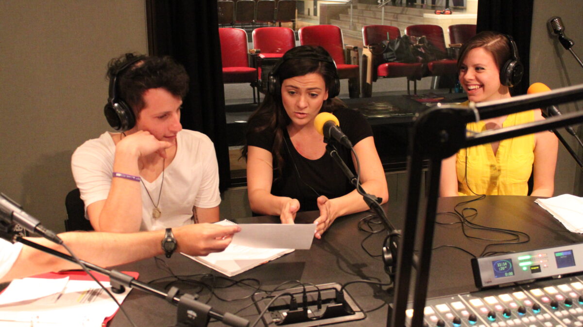 IMG_1328 Three adults wearing headphones reading a piece of paper into a microphone in a radio studio