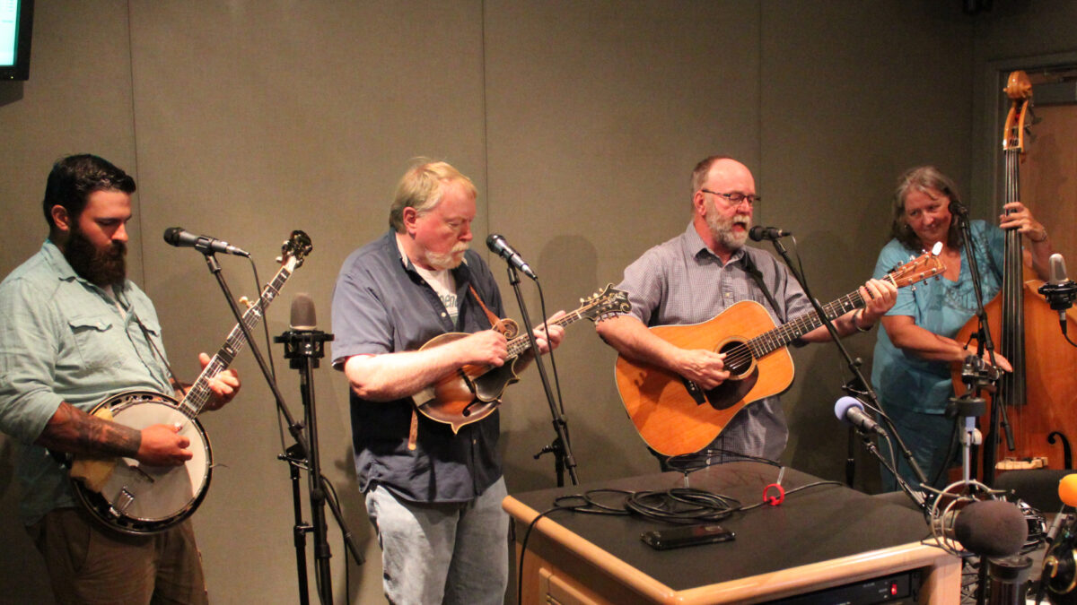 An adult playing banjo, an adult playing mandolin, an adult playing acoustic guitar and singing into a microphone, and an adult playing upright bass in a radio studio