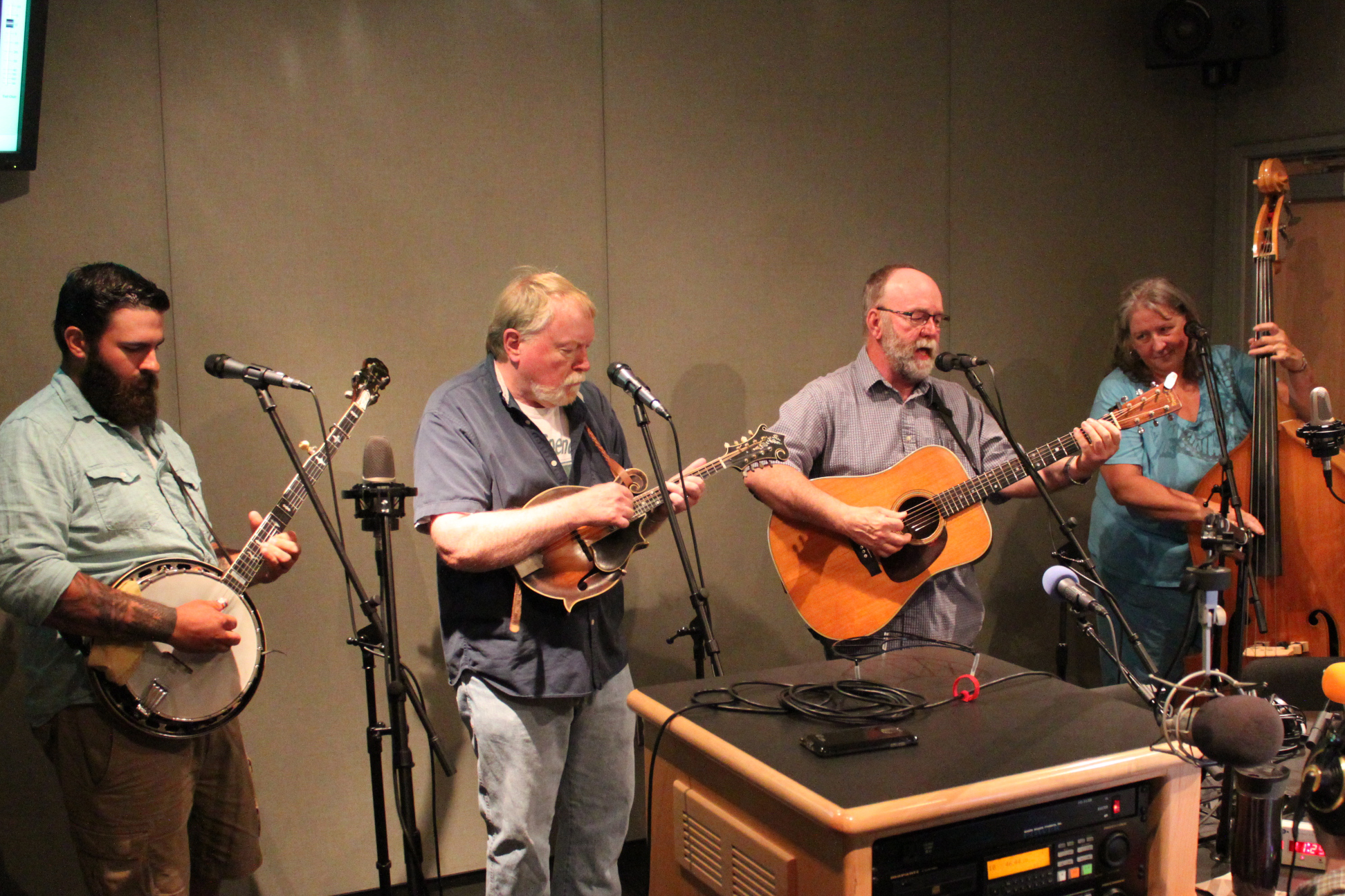 An adult playing banjo, an adult playing mandolin, an adult playing acoustic guitar and singing into a microphone, and an adult playing upright bass in a radio studio