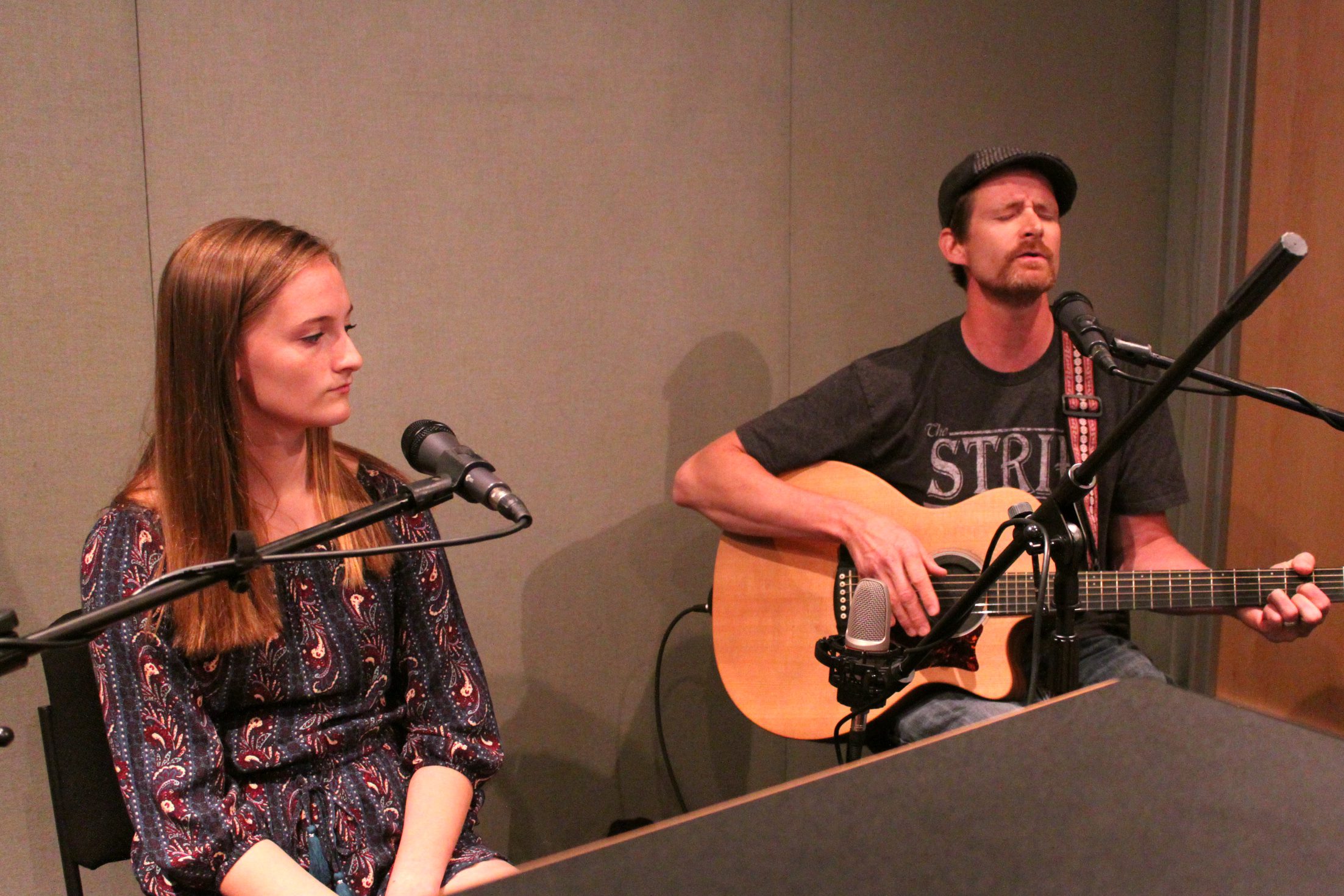A teen sitting behind a microphone and an adult playing an acoustic guitar sitting behind a microphone in a radio studio