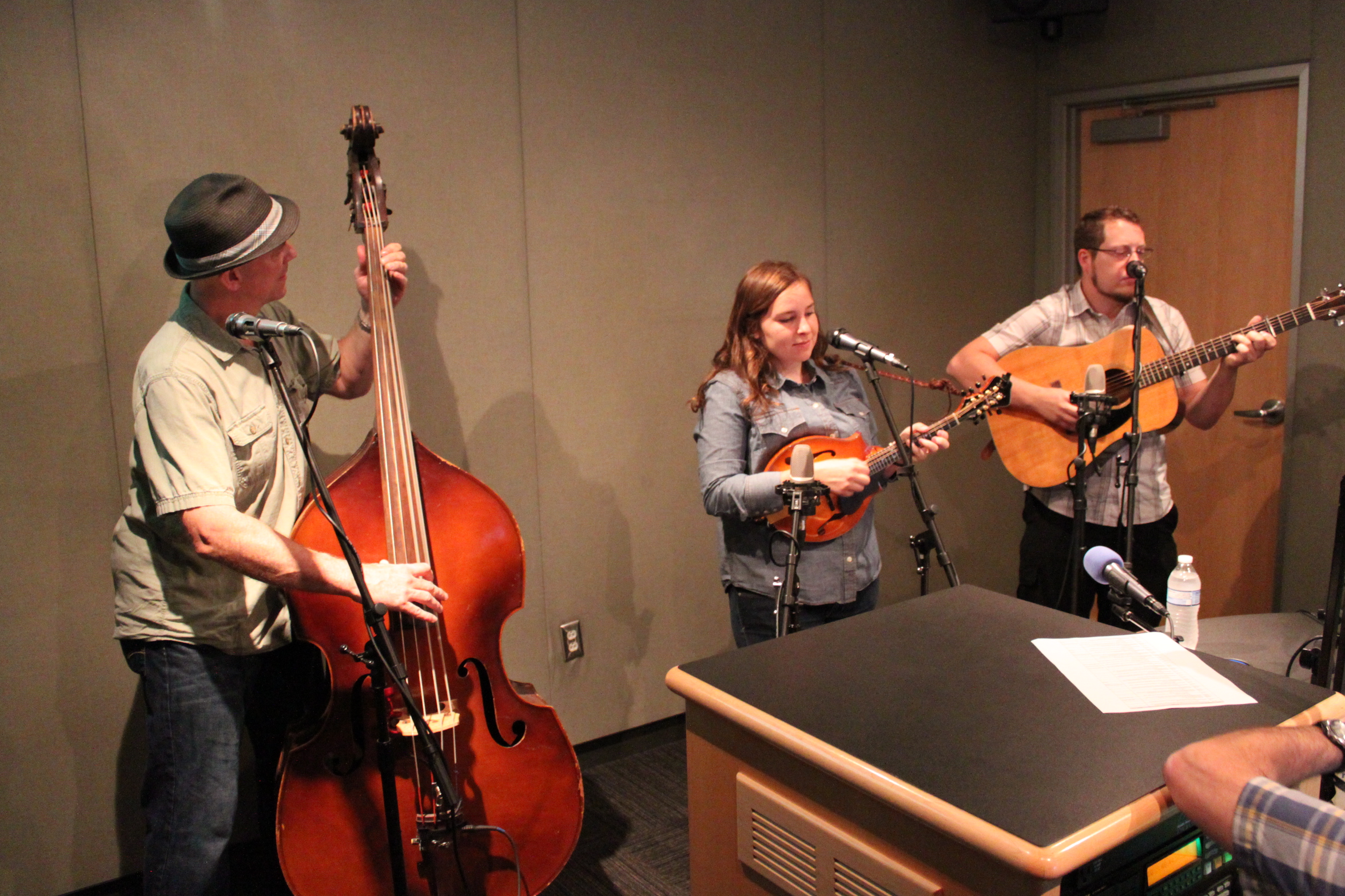 An adult playing an upright bass, an adult playing a mandolin, and an adult playing an acoustic guitar in a radio studio