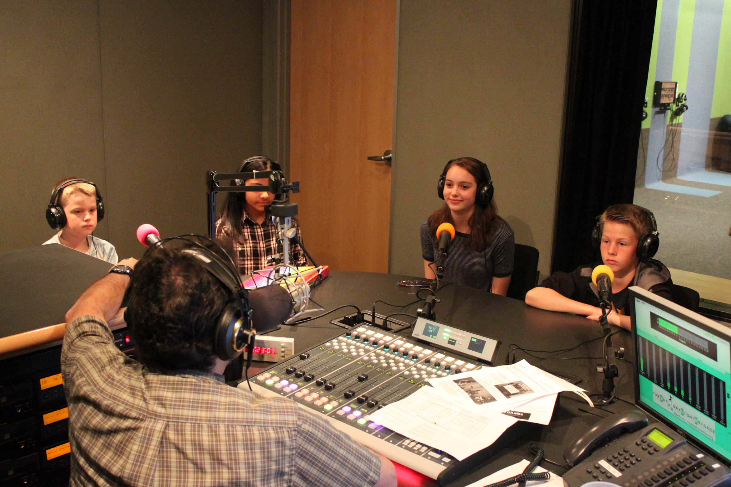 Four youth wearing headphones sitting behind microphones being interviewed by an adult wearing headphones sitting behind a microphone and a mixing board in a radio studio