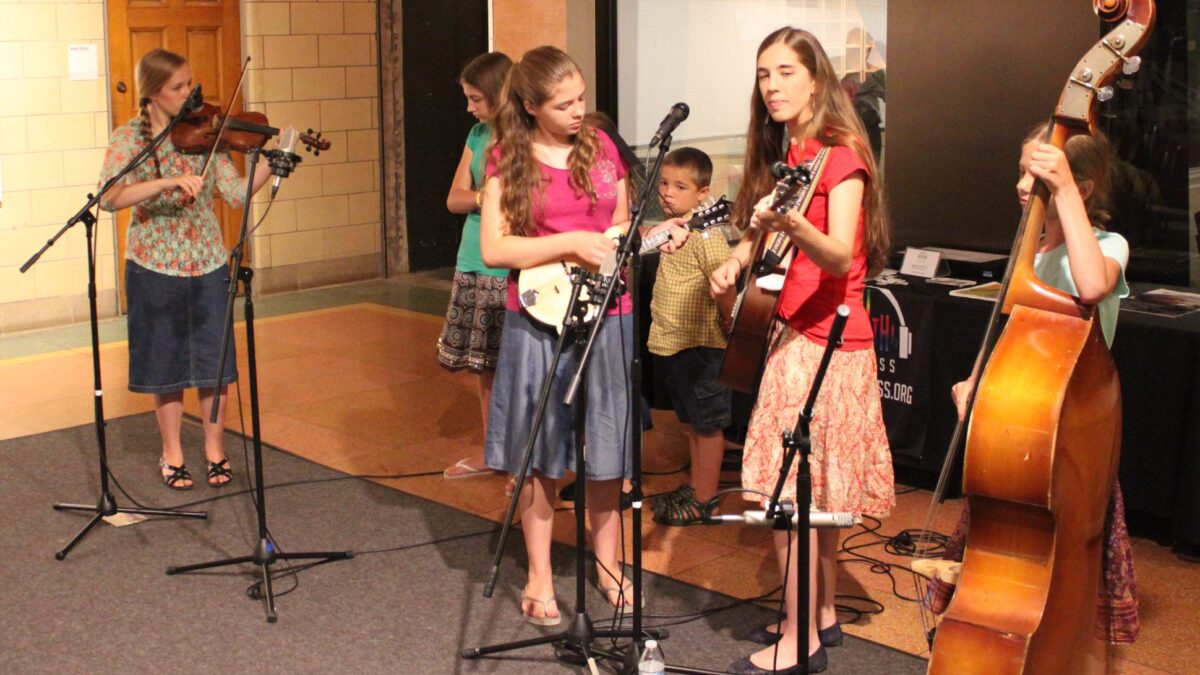 IMG_1462 A youth playing violin, three youth standing behind a microphone, a youth playing a mandolin, a youth playing an acoustic guitar, and a youth playing an upright bass
