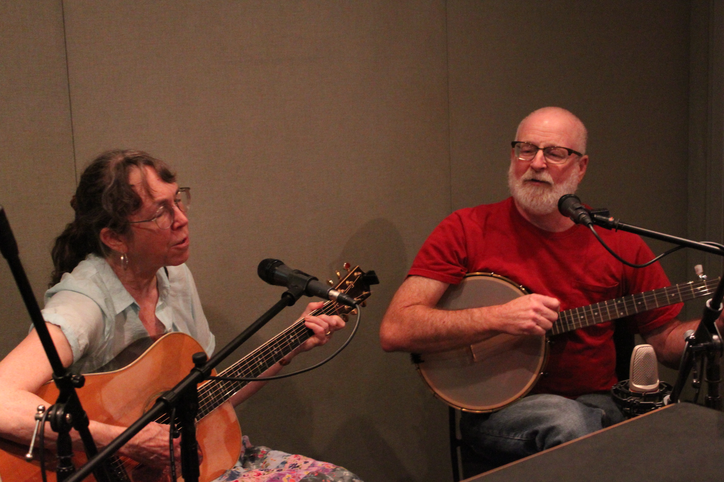 An adult playing an acoustic guitar and singing into a microphone and an adult playing a banjo and singing into a microphone in a radio studio