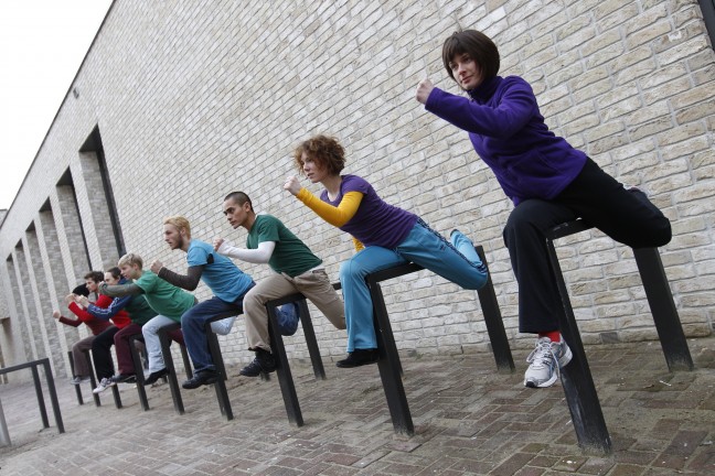 Eight adults making a leaping motion in front of a gray brick wall