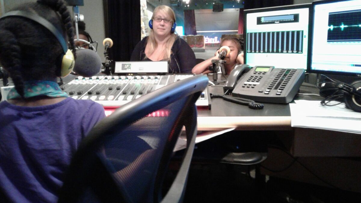An adult and two youth wearing headphones speaking into microphones and a youth wearing headphones sitting behind a microphone and a mixing board in a radio studio
