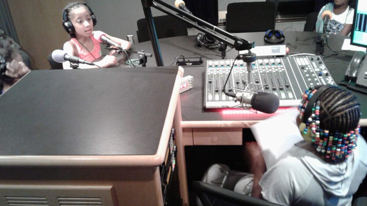 Three youth wearing headphones speaking into microphones and a youth sitting behind a microphone and a mixing board in a radio studio