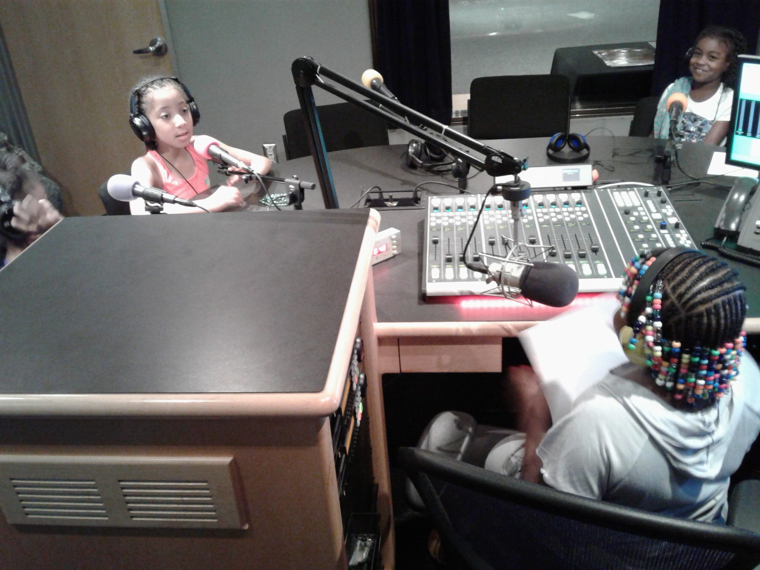 Three youth wearing headphones speaking into microphones and a youth sitting behind a microphone and a mixing board in a radio studio