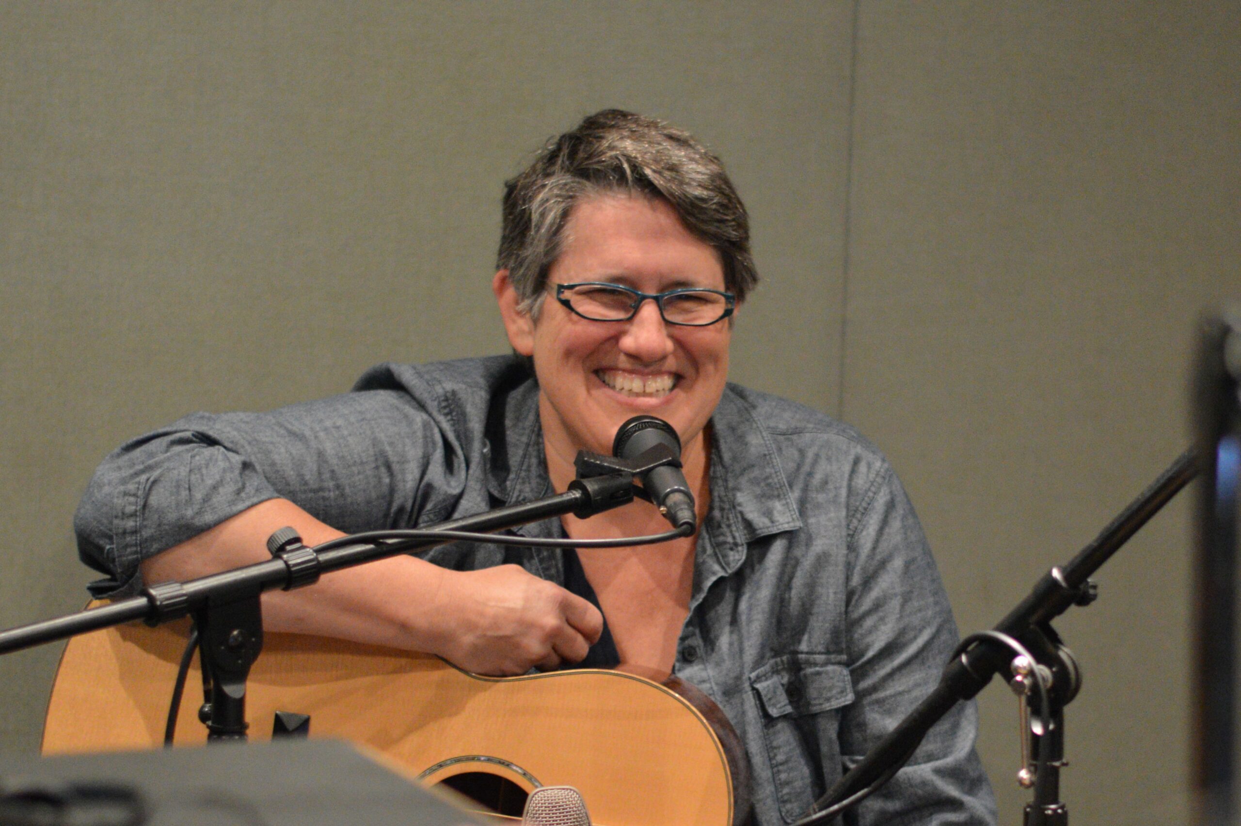 An adult holding a guitar smiling sitting behind a microphone in a radio studio