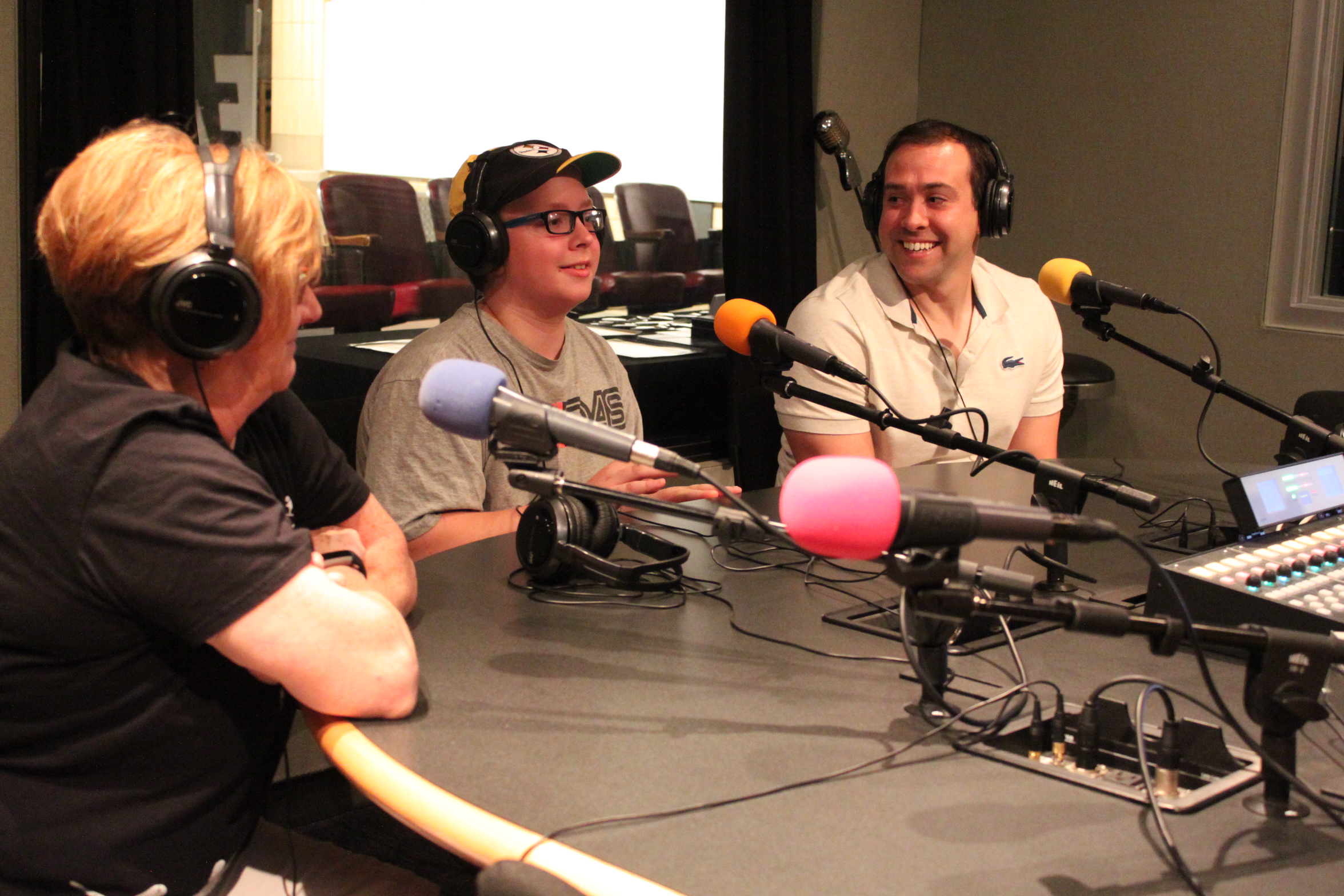 Two adults and a youth wearing headphones sitting behind microphones in a radio studio