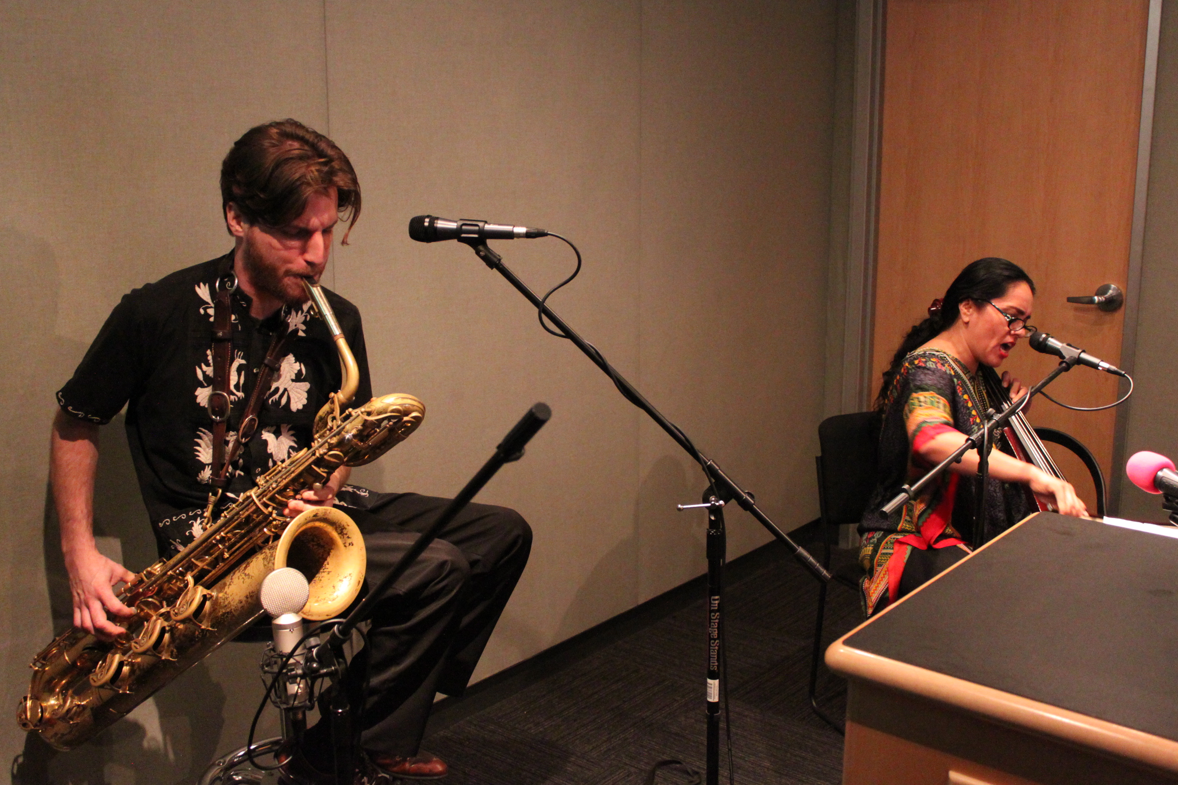 An adult playing a baritone saxophone and an adult playing a cello singing into a microphone in a radio studio
