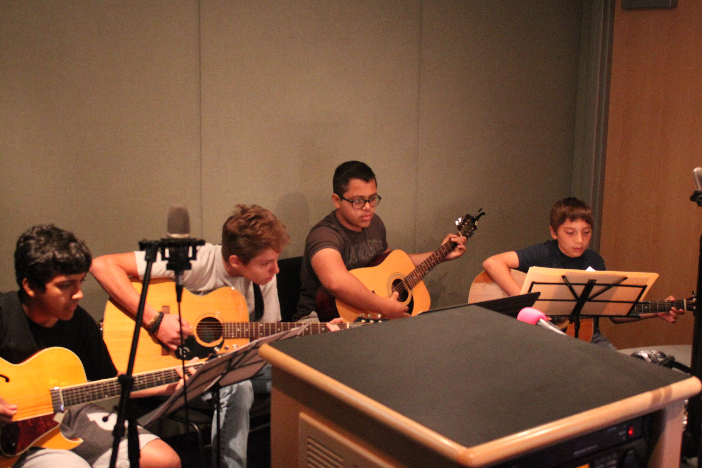 Four youth playing acoustic guitars looking at sheet music on music stands in a radio studio