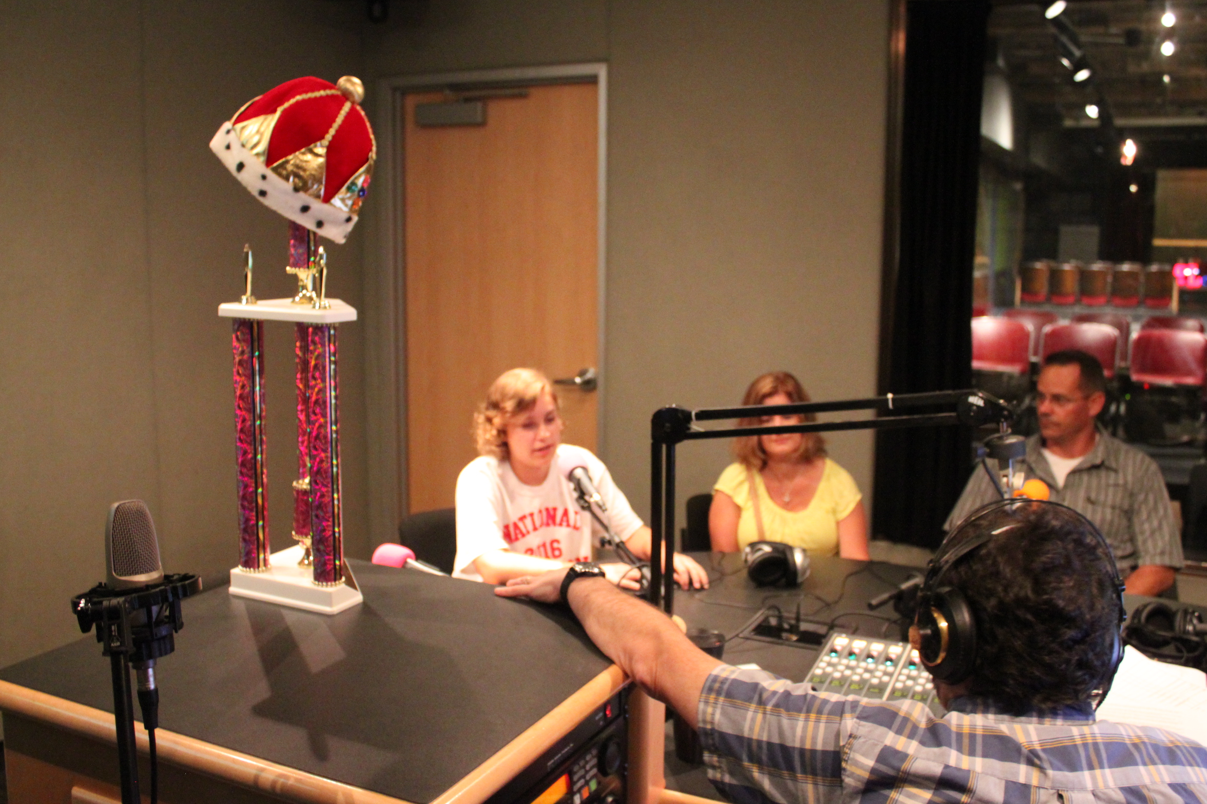 A teen and two adults speaking into microphones being interviewed by an adult wearing headphones sitting next to a large gold and red trophy in a radio studio