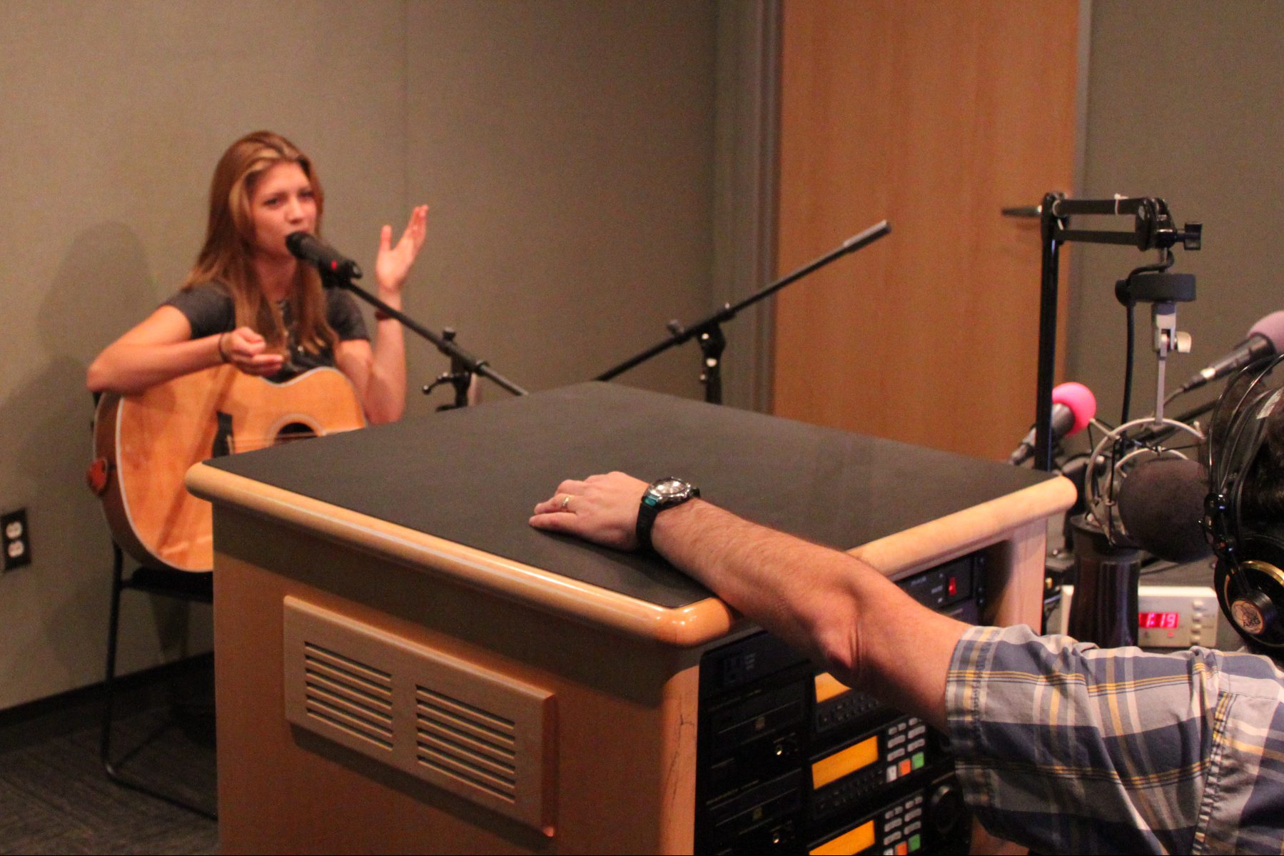 An adult holding an acoustic guitar speaking into a microphone being interviewed by an adult wearing headphones sitting behind a microphone in a radio studio