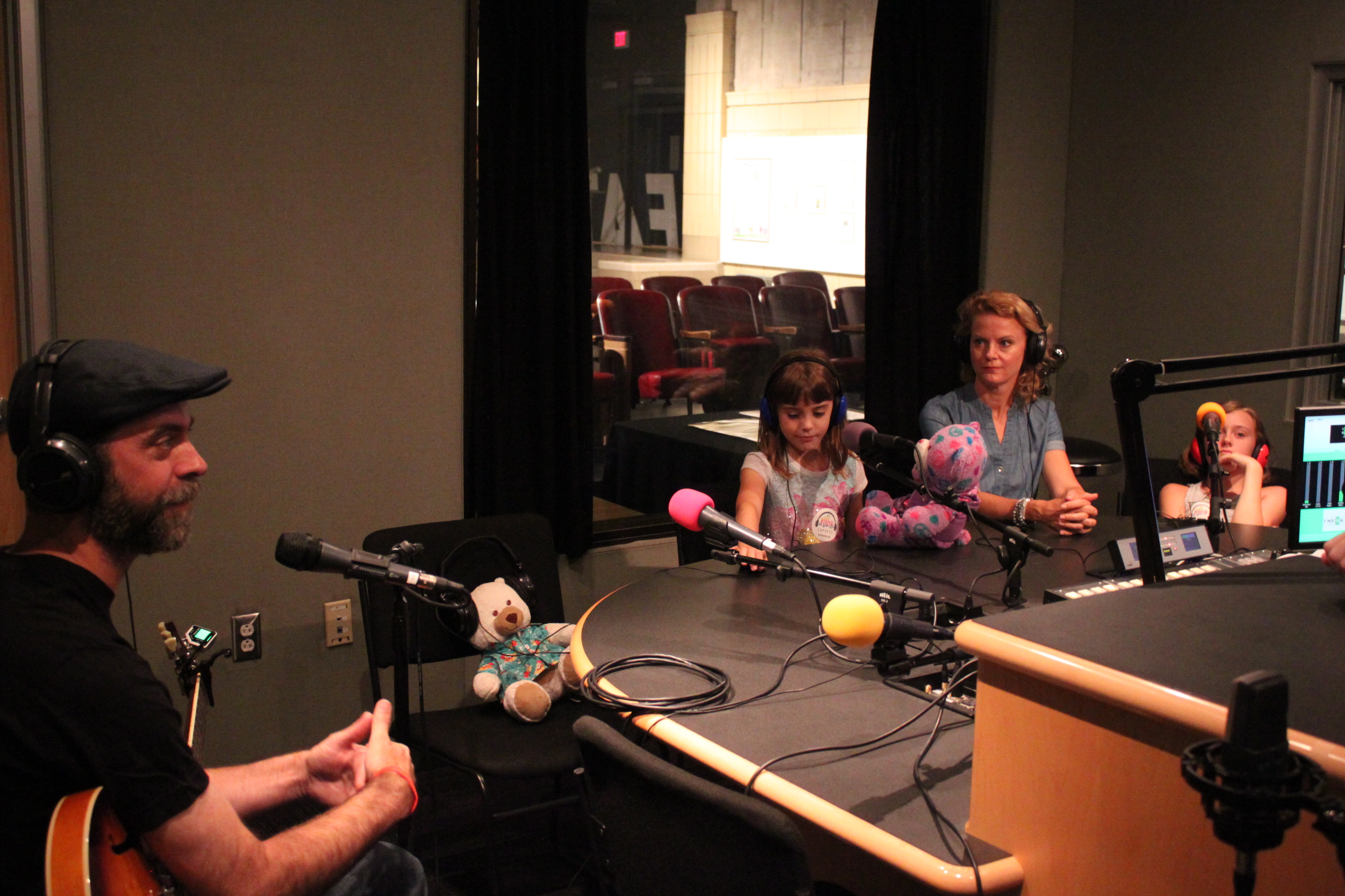 Two adults and two youth wearing headphones sitting behind microphones in a radio studio, one adult holding an acoustic guitar