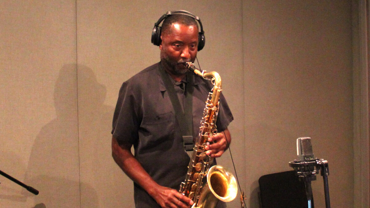 An adult playing a tenor saxophone into a microphone in a radio studio
