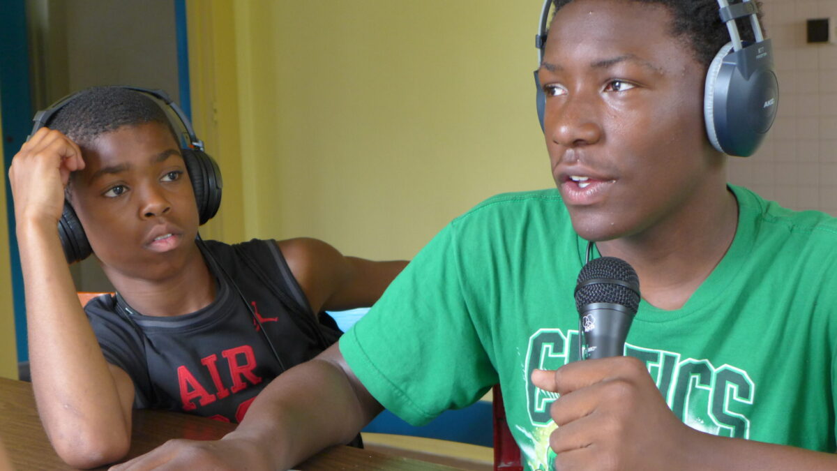 Two youth wearing headphones sitting around a brown table, one holding and speaking into a microphone
