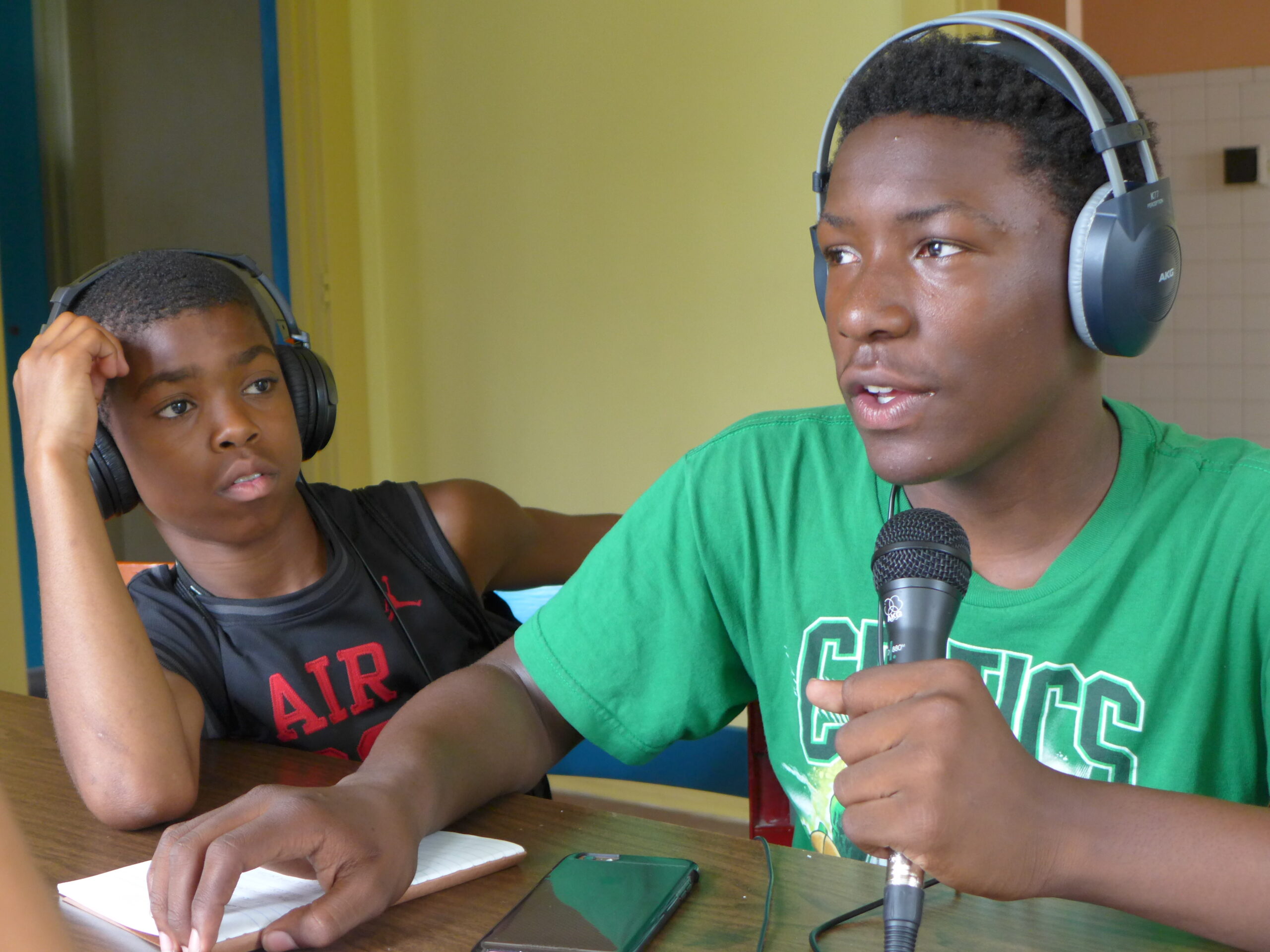 Two youth wearing headphones sitting around a brown table, one holding and speaking into a microphone