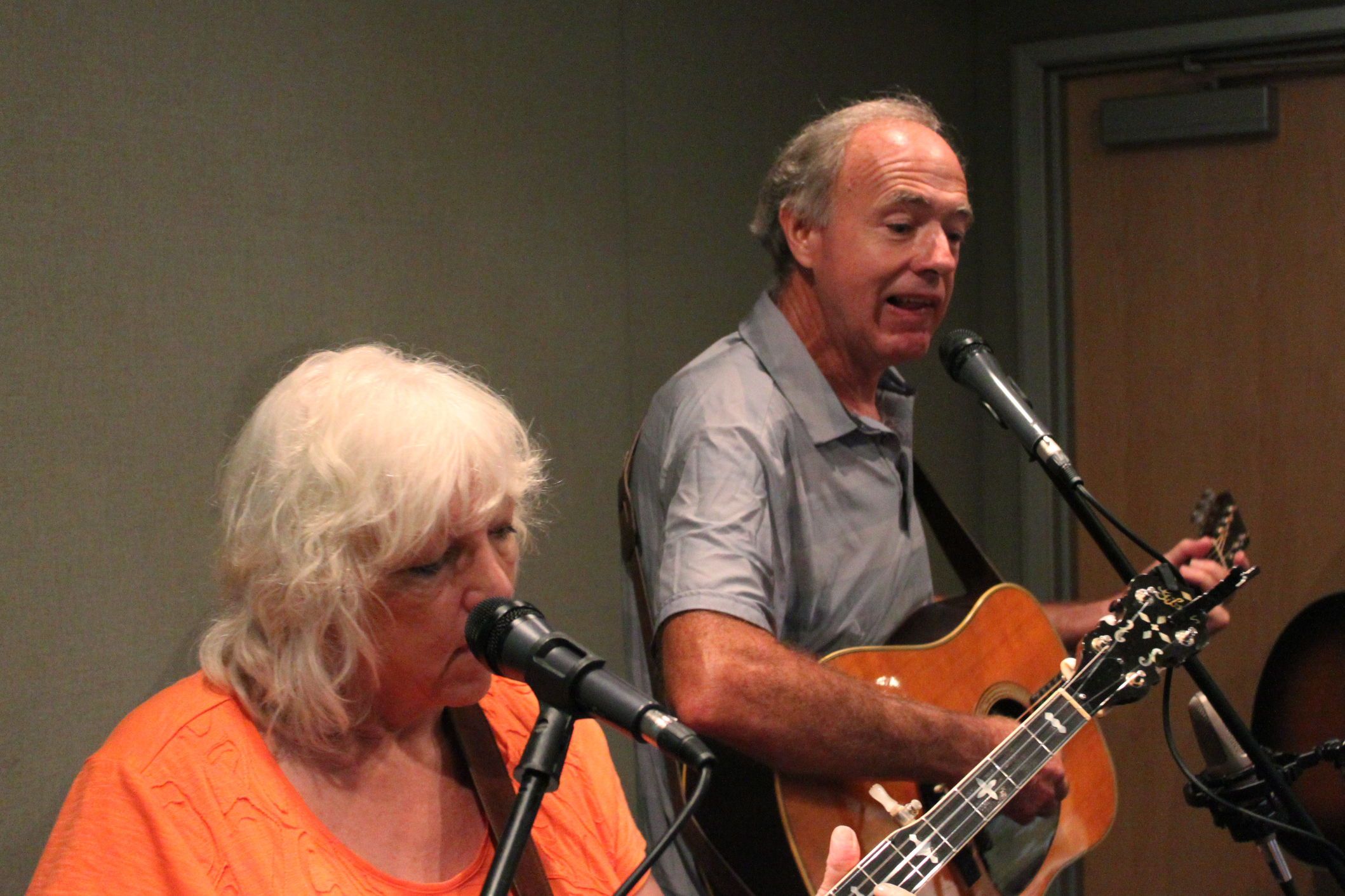 An adult singing into a microphone and an adult playing acoustic guitar singing into a microphone in a radio studio
