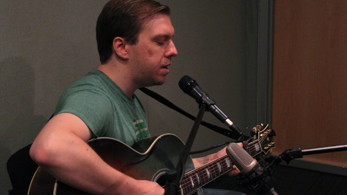 An adult playing acoustic guitar singing into a microphone in a radio studio