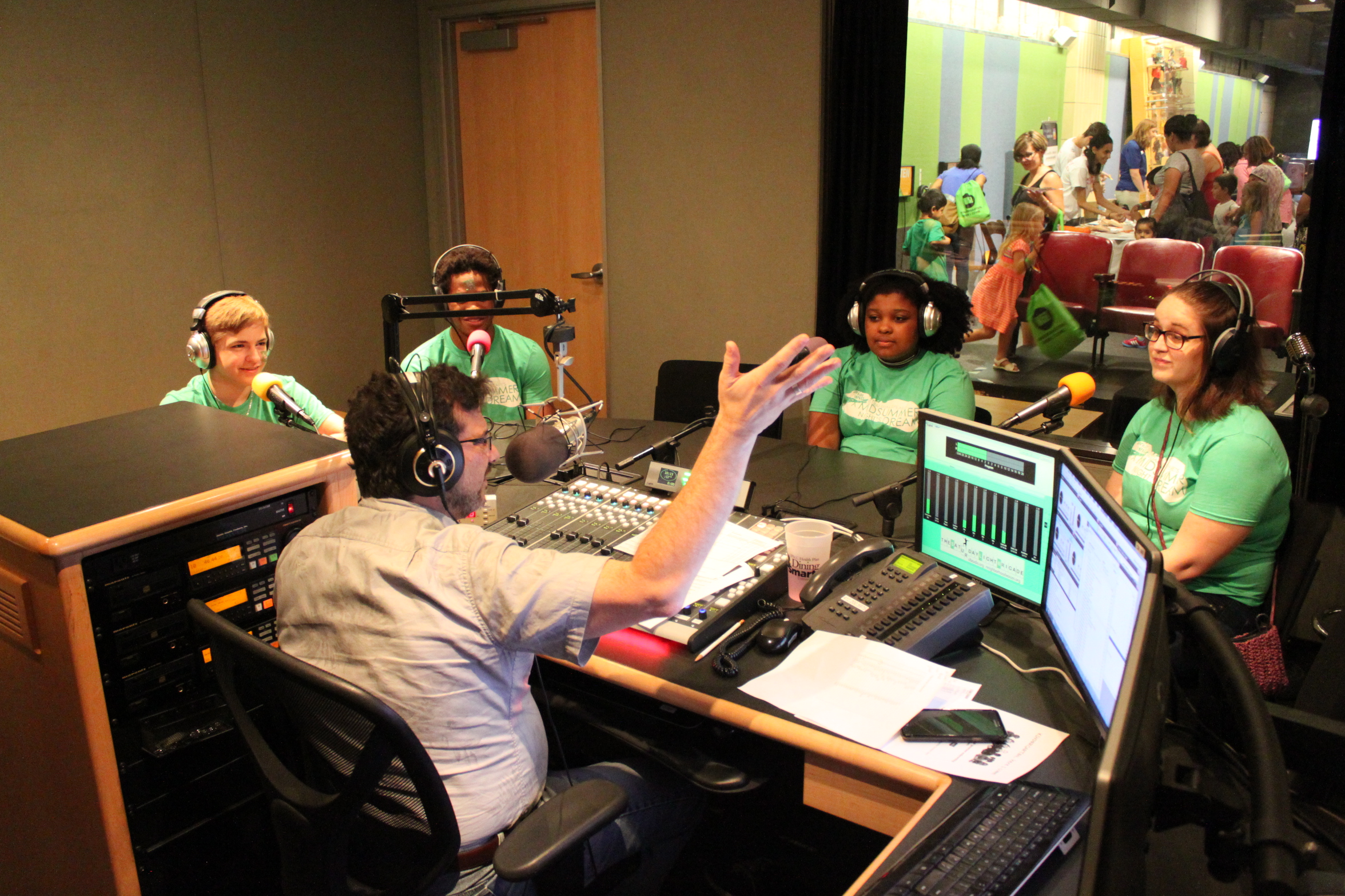 Four teens wearing headphones sitting behind microphones being interviewed by an adult wearing headphones sitting behind a microphone and a mixing board in a radio studio