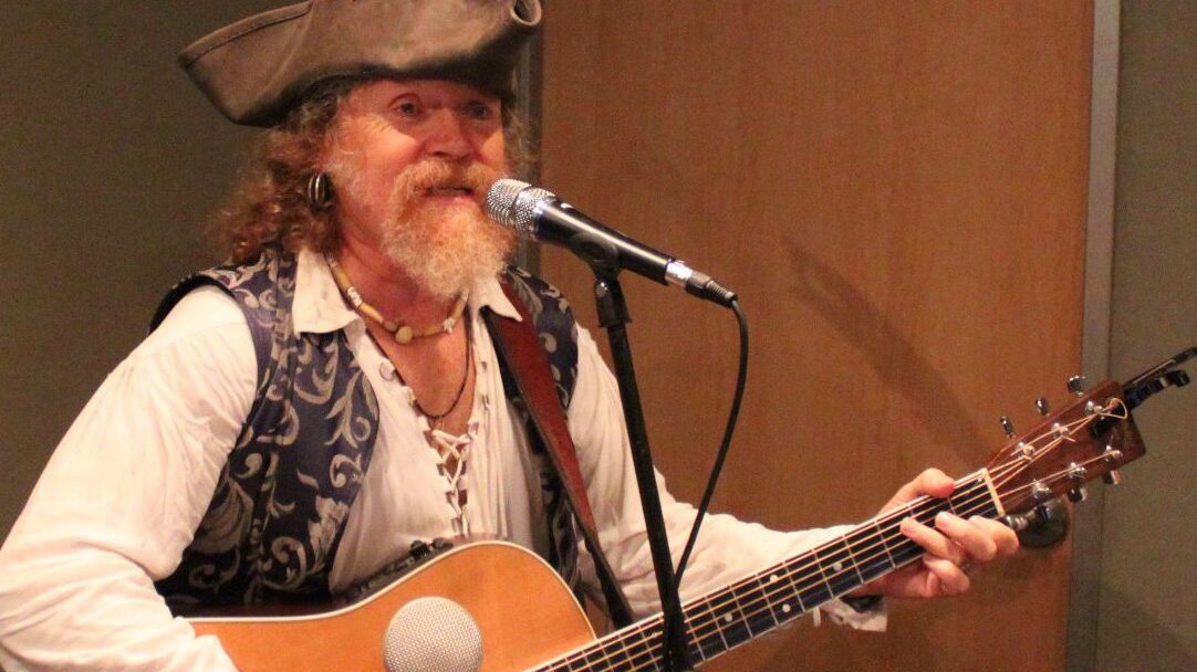 IMG_1914 An adult wearing pirate garb playing acoustic guitar and singing into a microphone in a radio studio