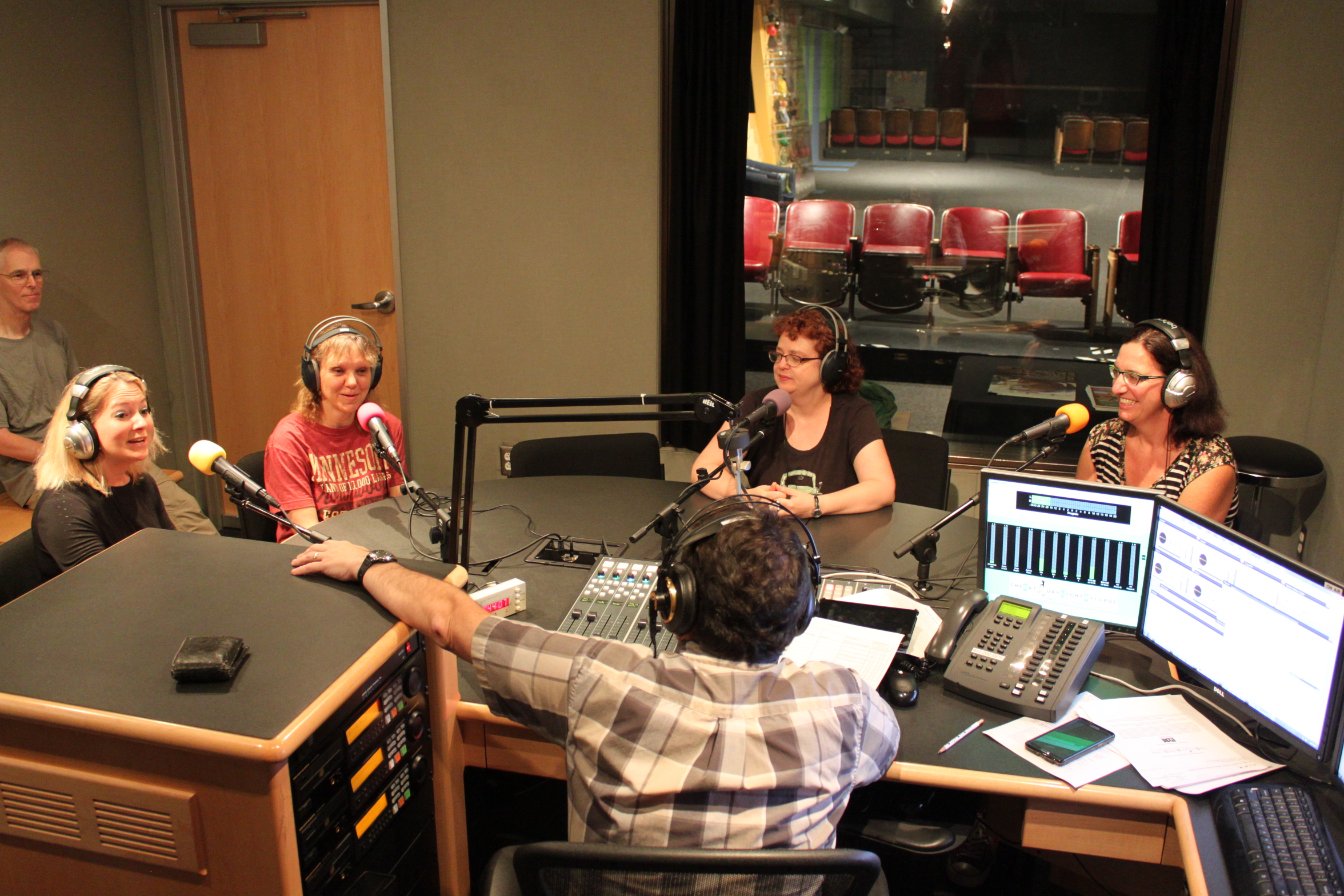 Four adults wearing headphones sitting behind microphones and one adult sitting in the background being interviewed by an adult wearing headphones sitting behind a microphone and a mixing board in a radio studio