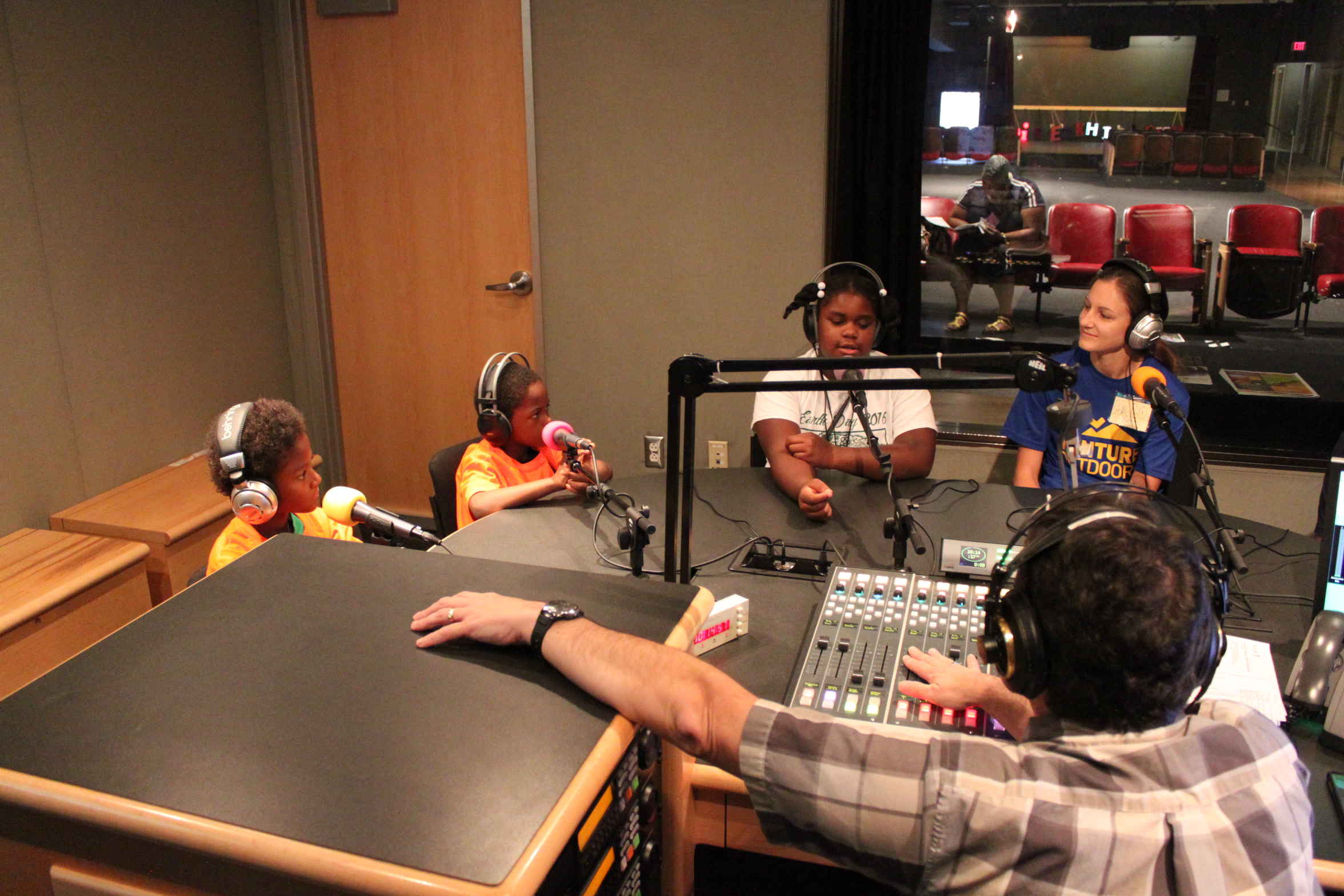 Three youth and an adult wearing headphones speaking into microphones being interviewed by an adult wearing headphones sitting behind a microphone and a mixing board in a radio studio