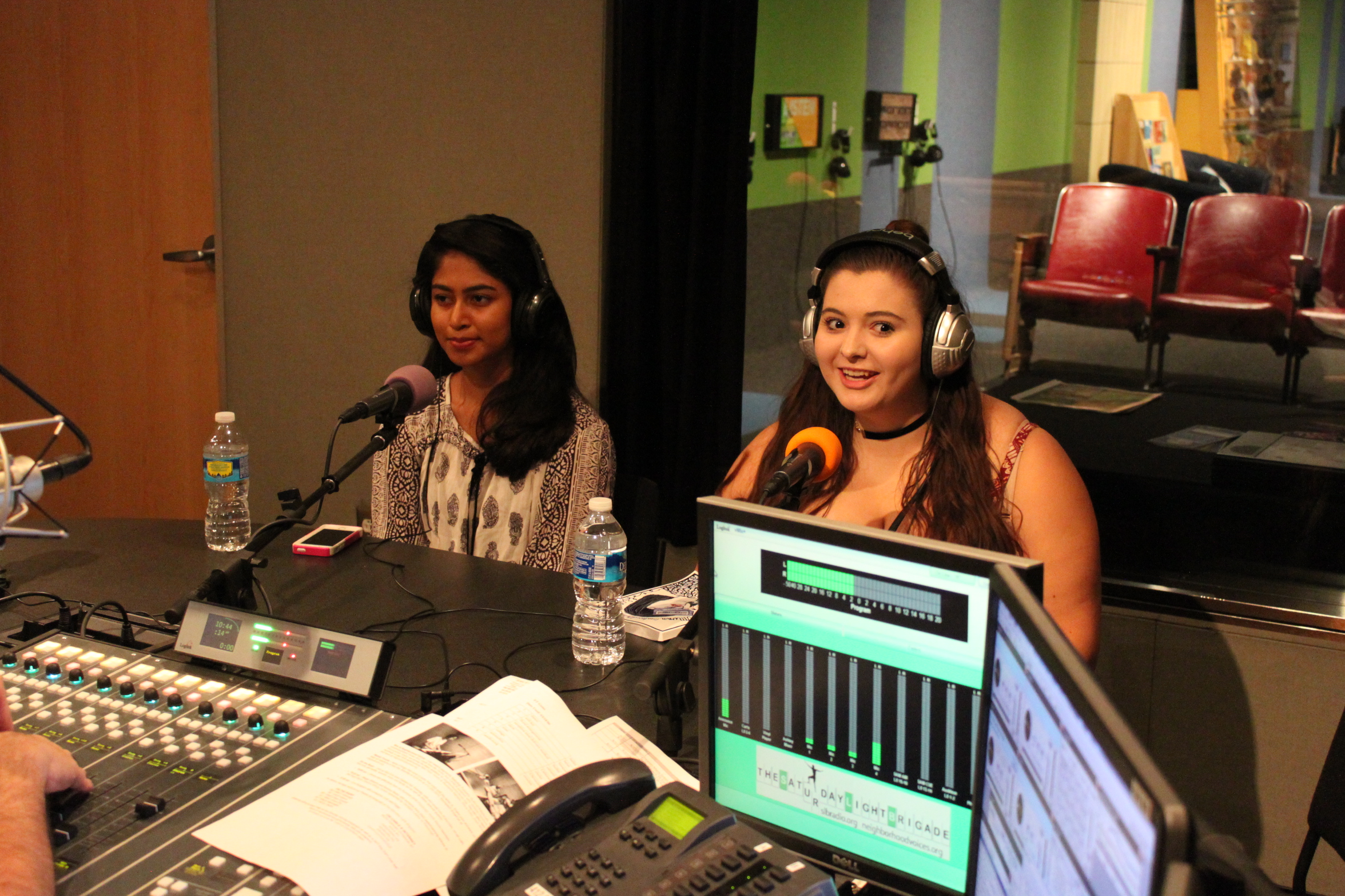 Two teens wearing headphones smiling while sitting behind microphones in a radio studio