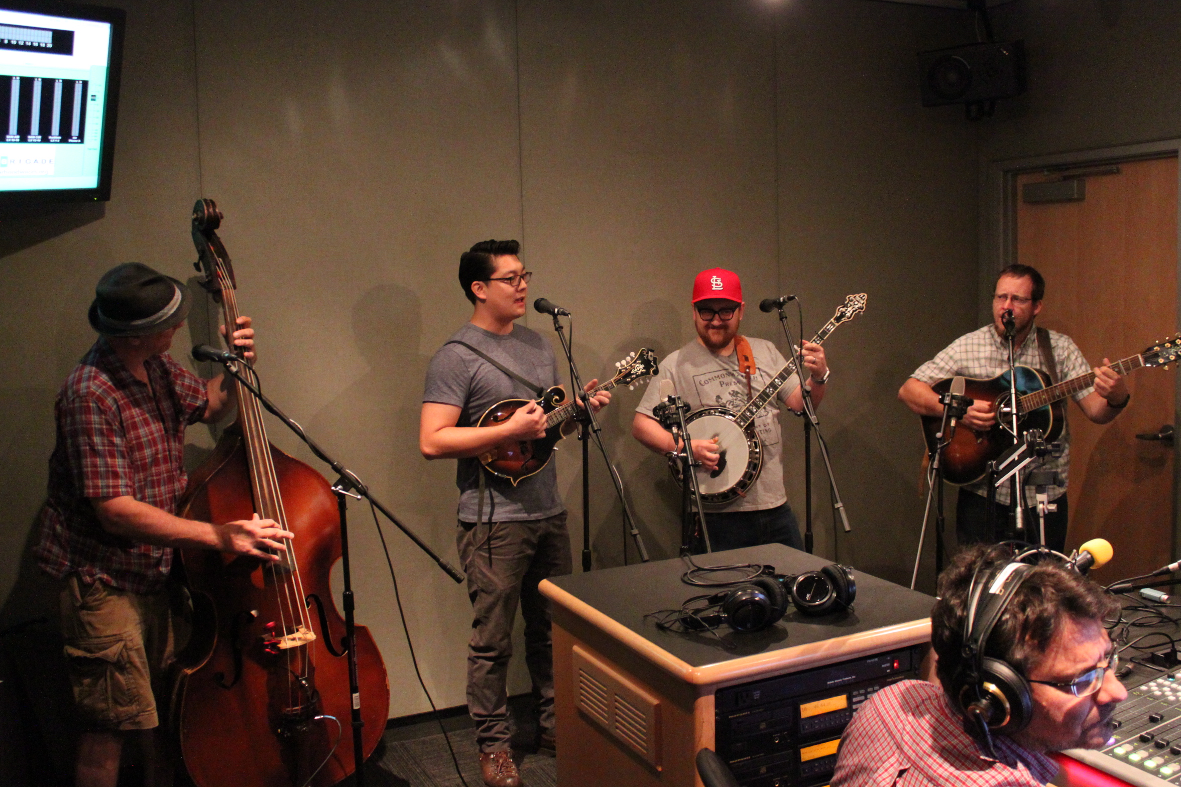 An adult playing upright bass, an adult playing mandolin, an adult playing banjo, and an adult playing acoustic guitars, all standing behind microphones as an adult wearing headphones sitting behind a microphone listens in a radio studio