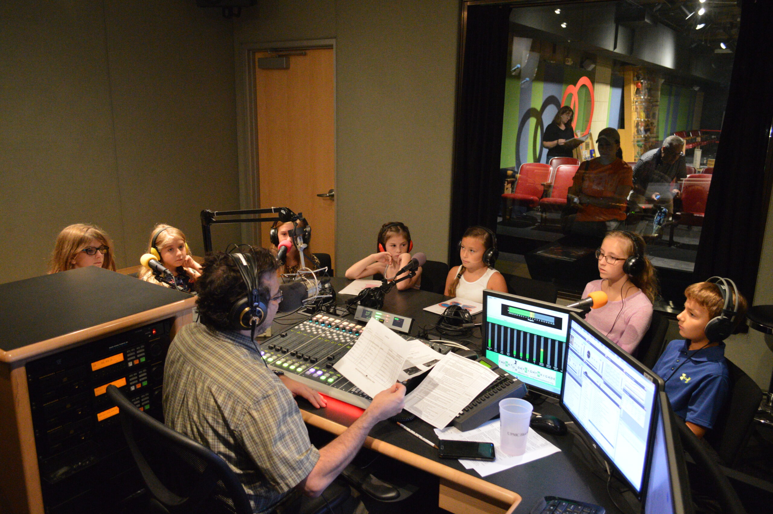 Six youth and one adult sitting behind microphones being interviewed by an adult wearing headphones sitting behind a microphone and a mixing board in a radio studio