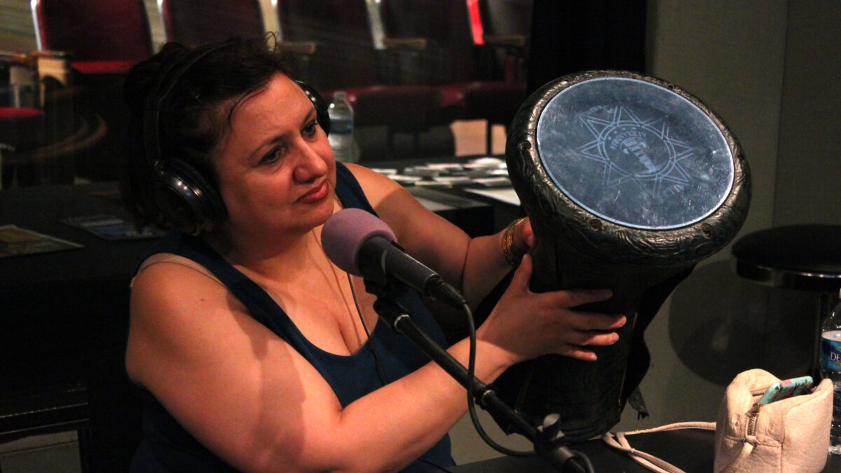 An adult wearing headphones sitting behind a microphone holding a percussive instrument in a radio studio