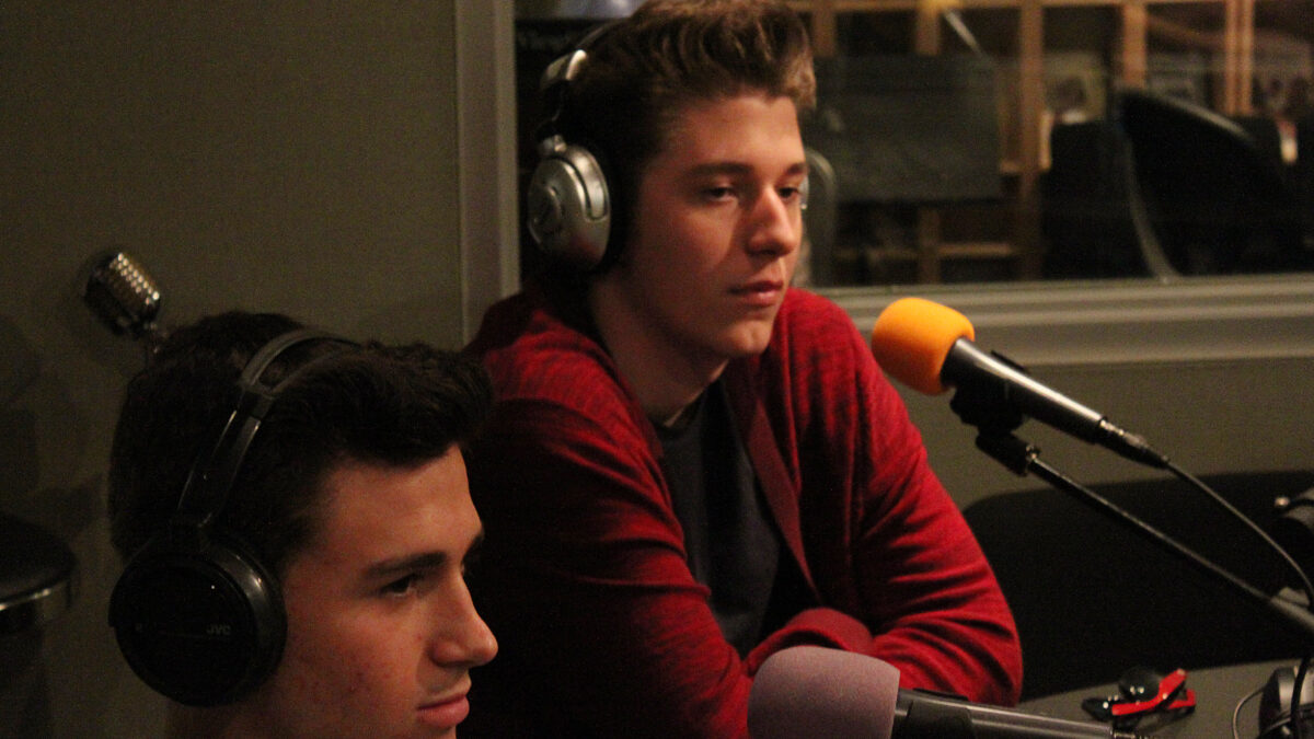 Two teens wearing headphones sitting behind microphones in a radio studio