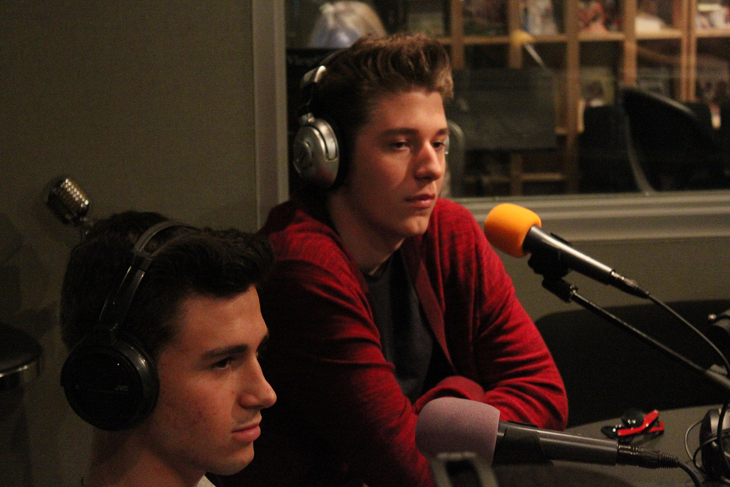 Two teens wearing headphones sitting behind microphones in a radio studio