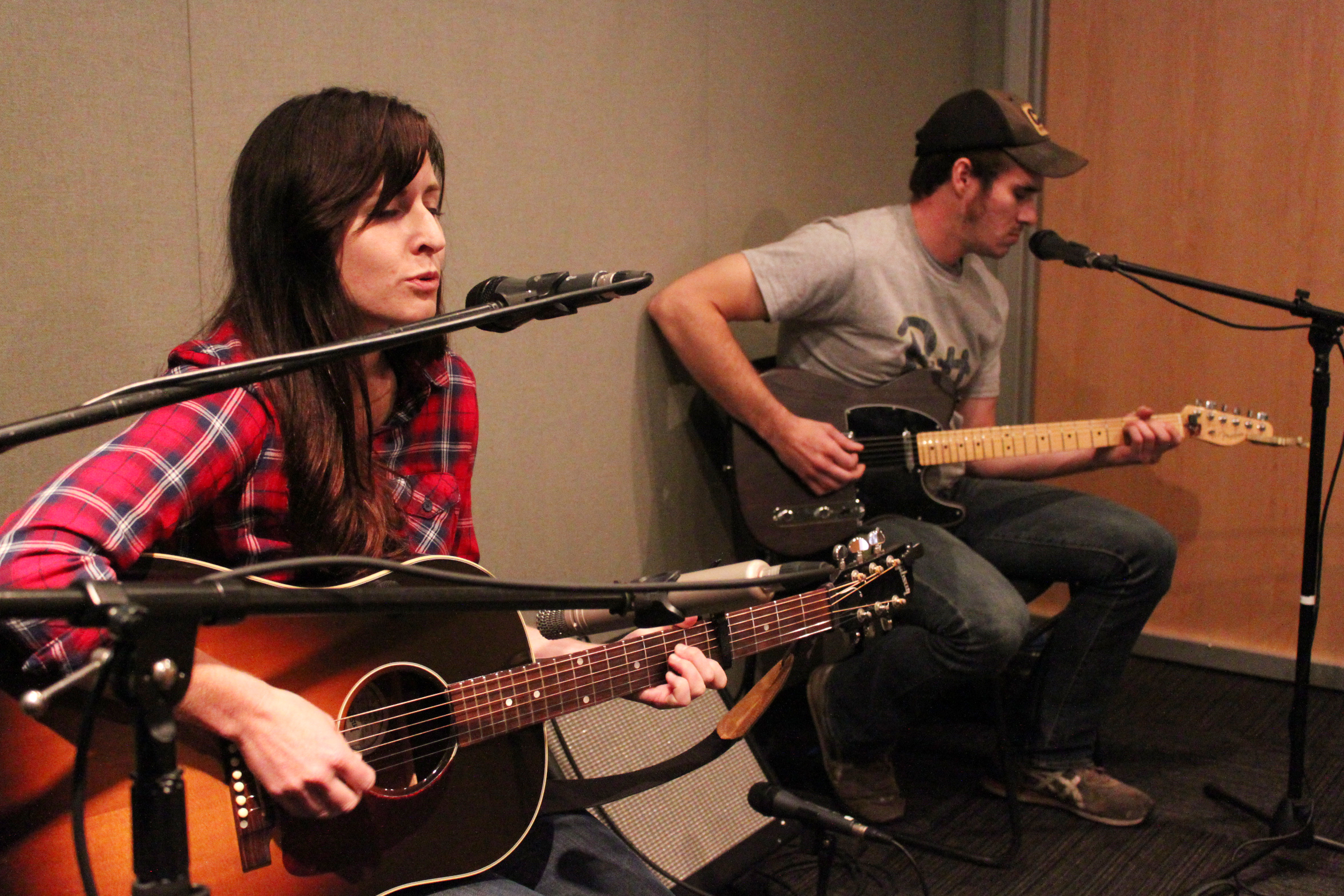 An adult playing acoustic guitar and singing into a microphone and an adult playing electric guitar in a radio studio
