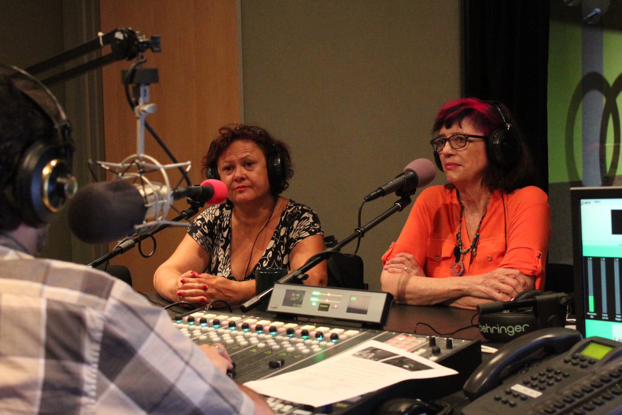 Two adults wearing headphones speaking into microphones being interviewed by an adult wearing headphones sitting behind a microphone and a mixing board in a radio studio