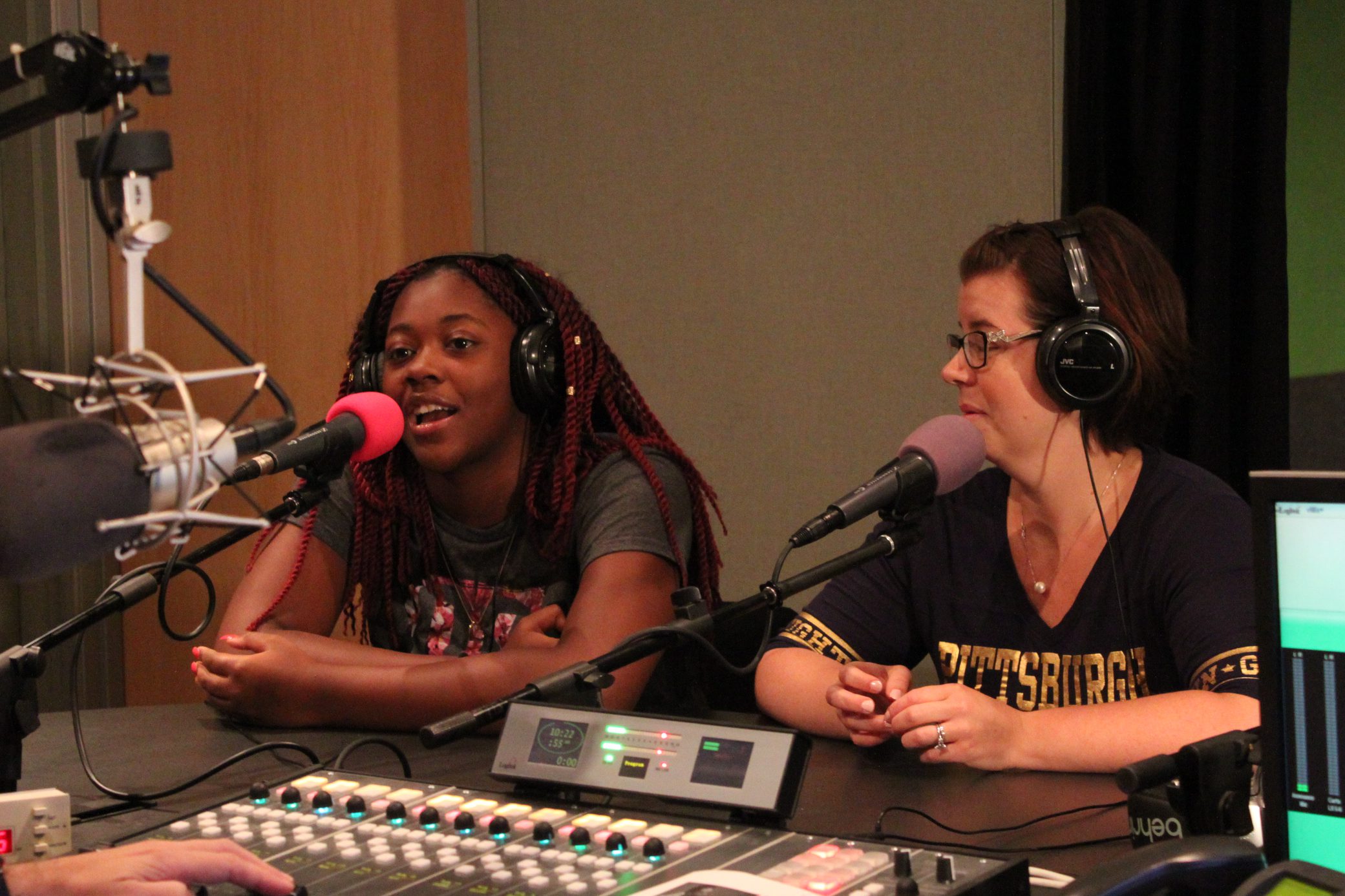 A teen and an adult wearing headphones speaking into microphones in a radio studio