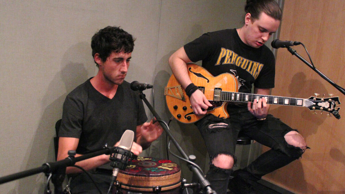An adult playing an electric guitar and an adult playing a conga in a radio studio