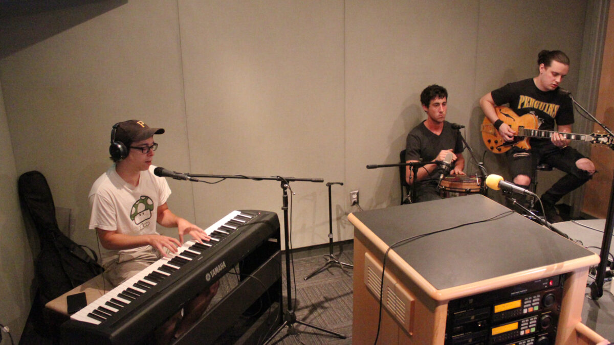 An adult wearing headphones playing a piano keyboard and singing into a microphone, an adult playing a conga, and an adult playing an electric guitar in a radio studio