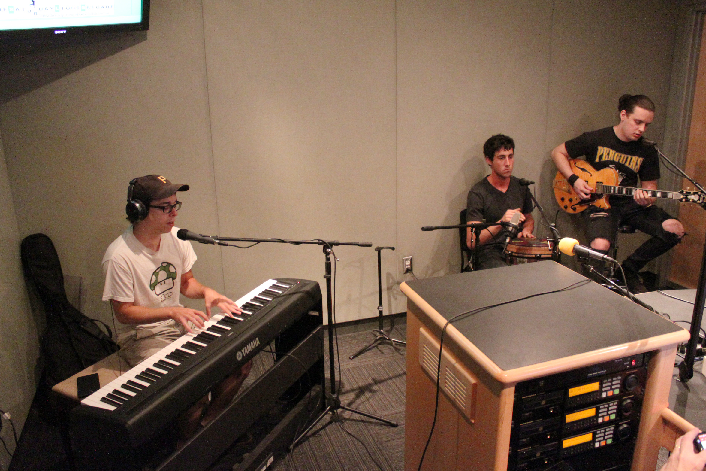 An adult wearing headphones playing a piano keyboard and singing into a microphone, an adult playing a conga, and an adult playing an electric guitar in a radio studio