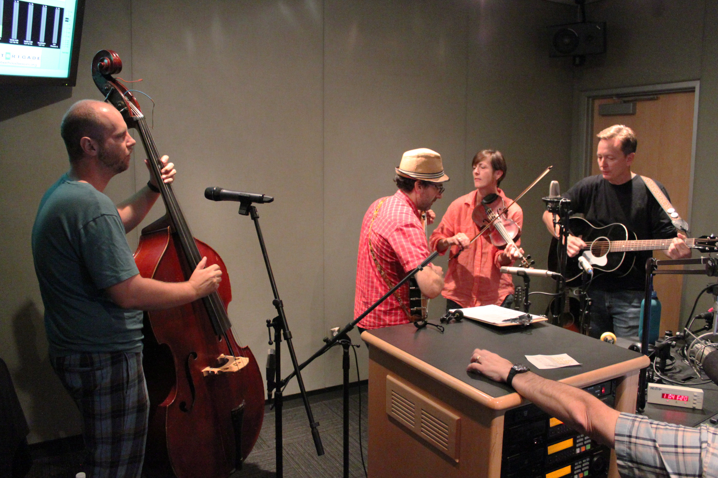 An adult playing upright bass, an adult playing banjo, an adult playing a fiddle, and an adult playing acoustic guitar in a radio studio