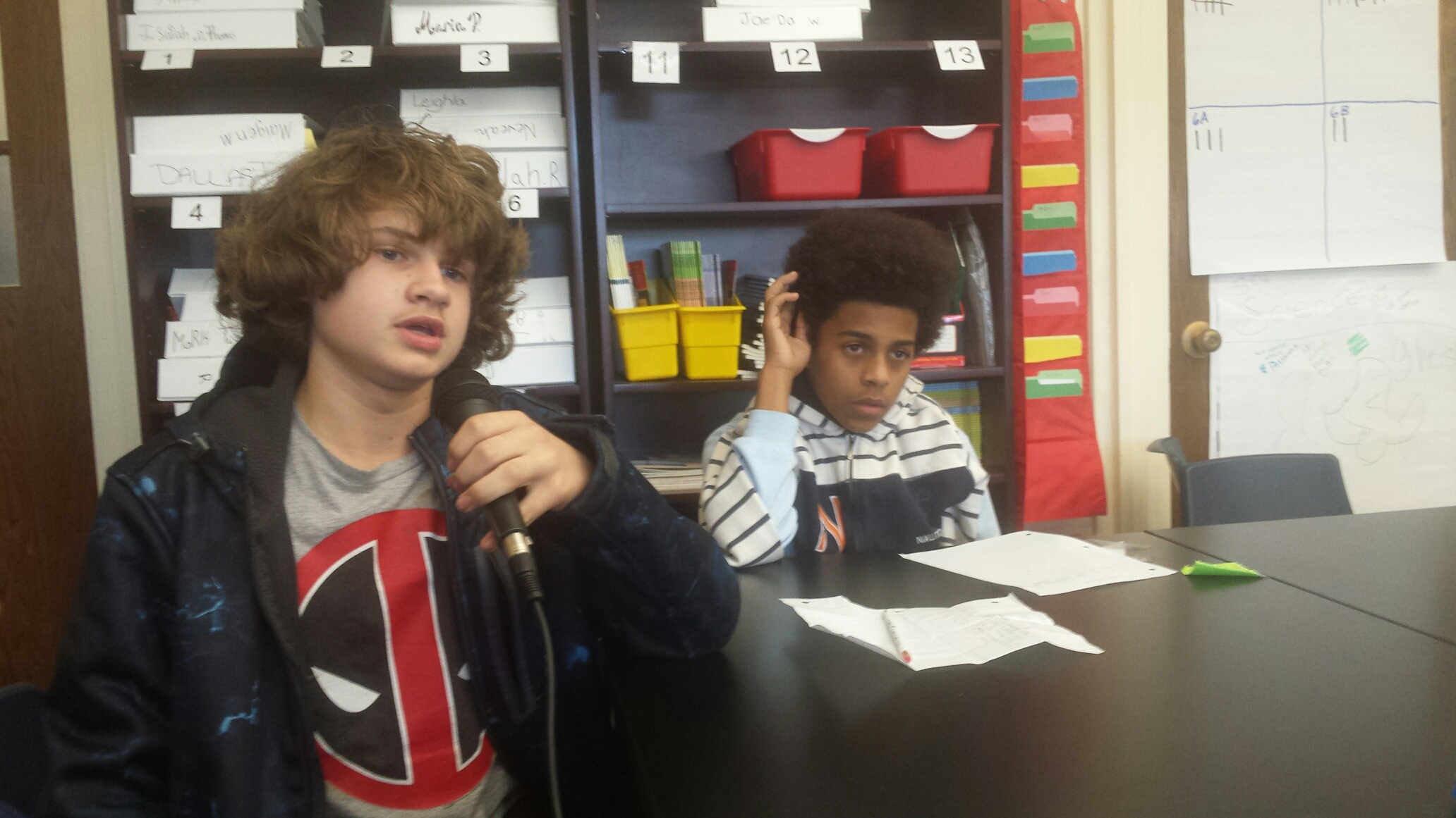 Two youth sitting around pieces of paper on a black table, one holding and speaking into a microphone