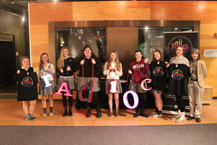 Eight teens and an adult smiling and posing in profile, three of the teens holding t-shirts and the other four teens holding letters