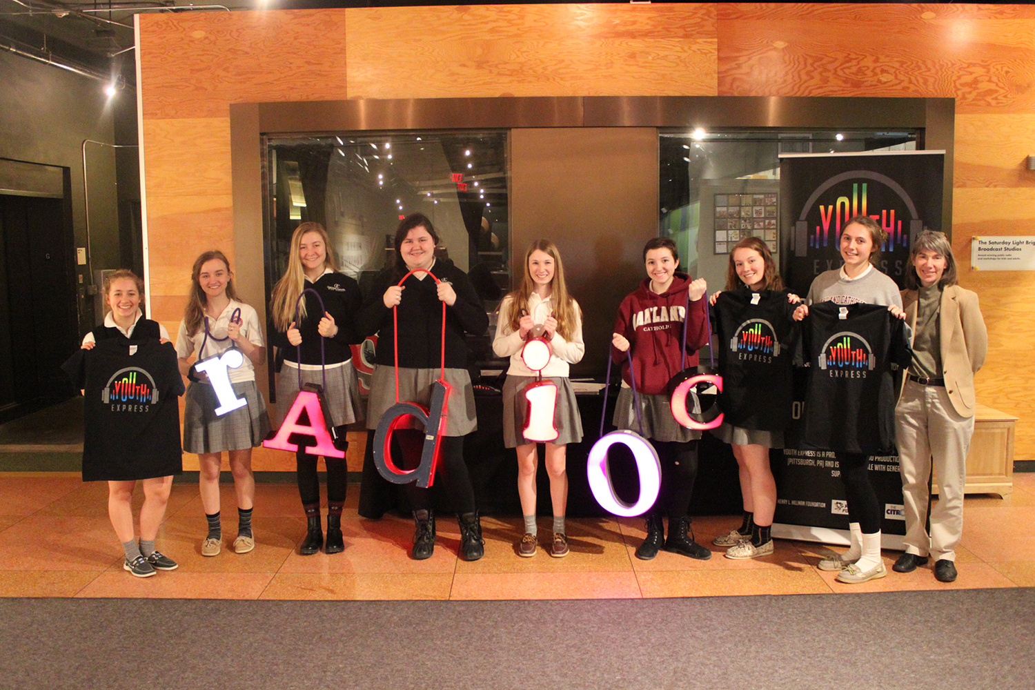 Eight teens and an adult smiling and posing in profile, three of the teens holding t-shirts and the other four teens holding letters