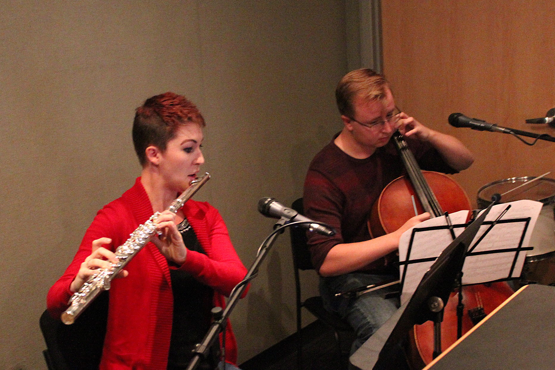 An adult playing a flute and an adult playing a cello, both looking at sheet music on music stands in a radio studio