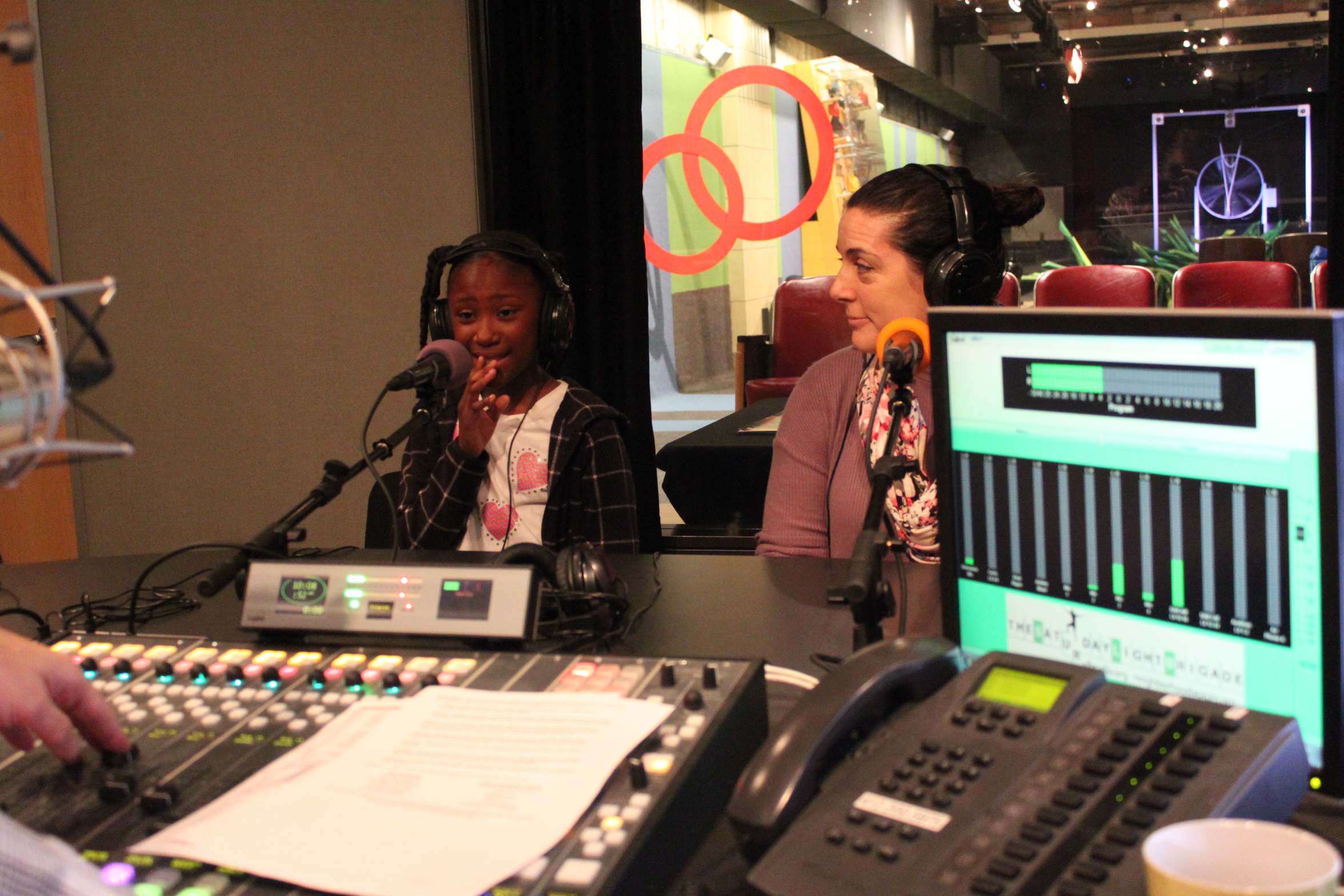 A youth and an adult wearing headphones speaking into microphones in a radio studio
